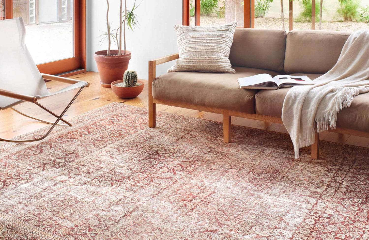 Bright living room with a distressed Persian-style rug in faded red, beige, and subtle blue tones, paired with a light wood sofa, indoor plants, and sunlit windows.