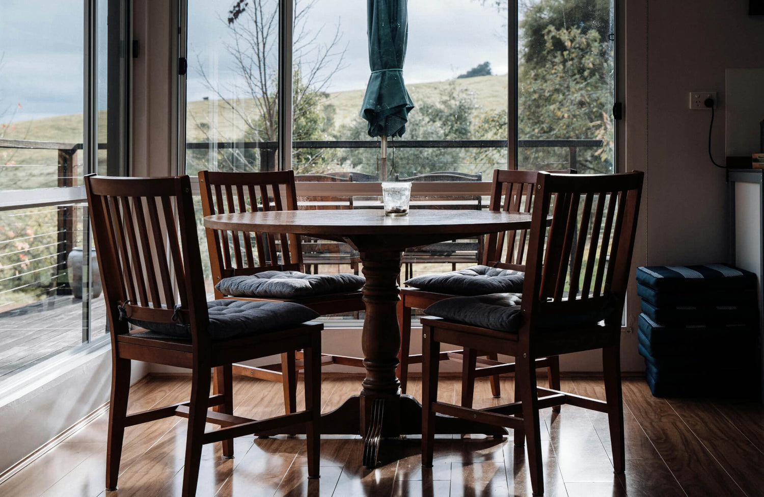 A warm, sunlit dining nook features a wooden round dining table set with four cushioned chairs, framed by large glass doors that open to a serene outdoor view.