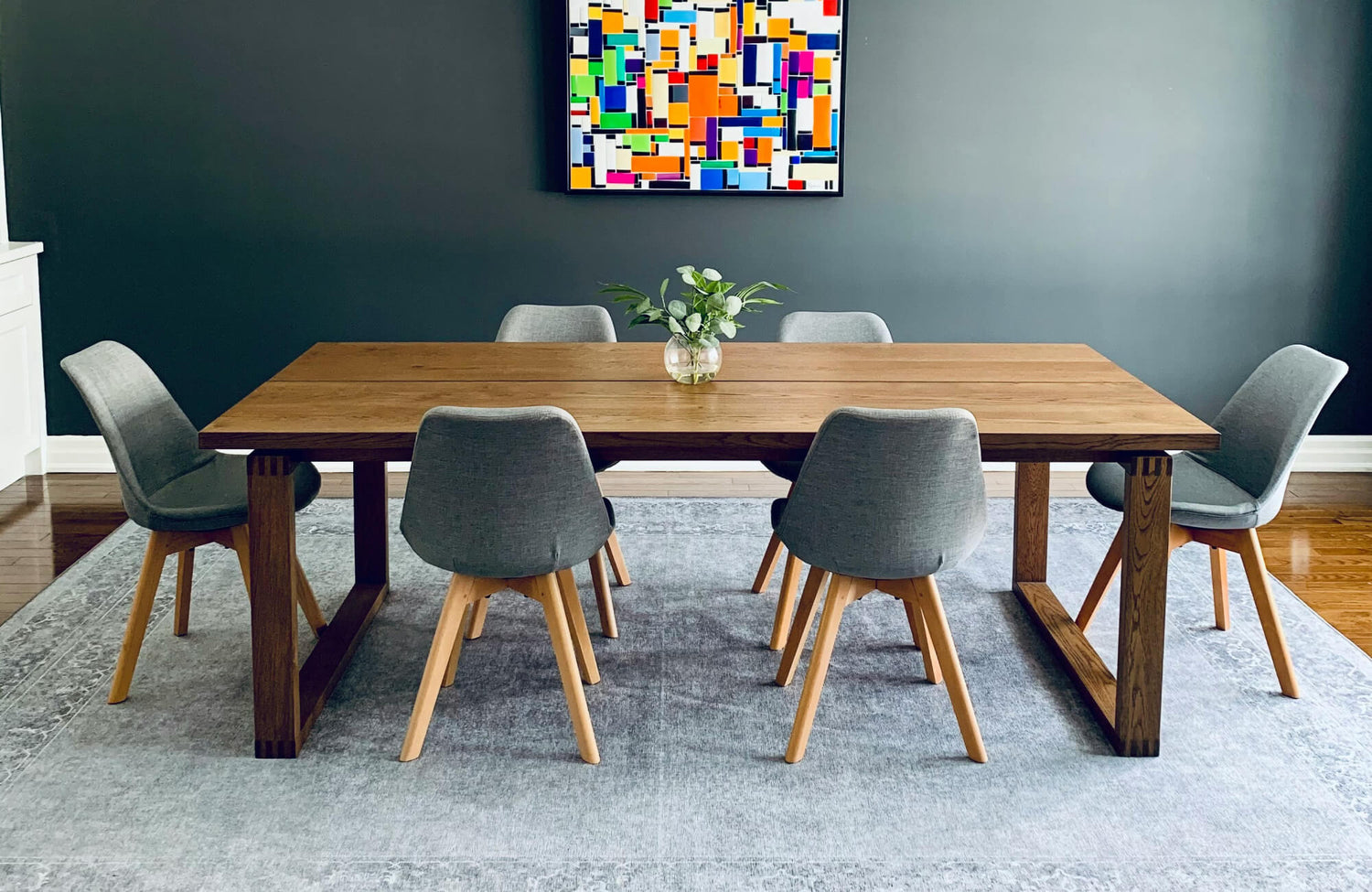 Minimalist dining room with a solid wood table, six grey fabric chairs, and a vibrant abstract painting on a dark wall.