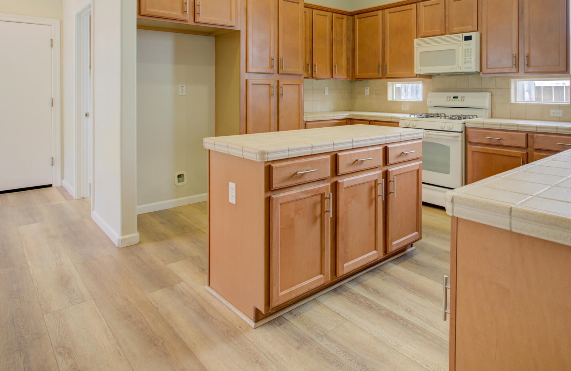 Warm, inviting kitchen featuring light wood-look flooring that flows seamlessly beneath the cabinetry and island, grounding the space with natural texture while complementing the honey-toned wood cabinets and soft neutral finishes.