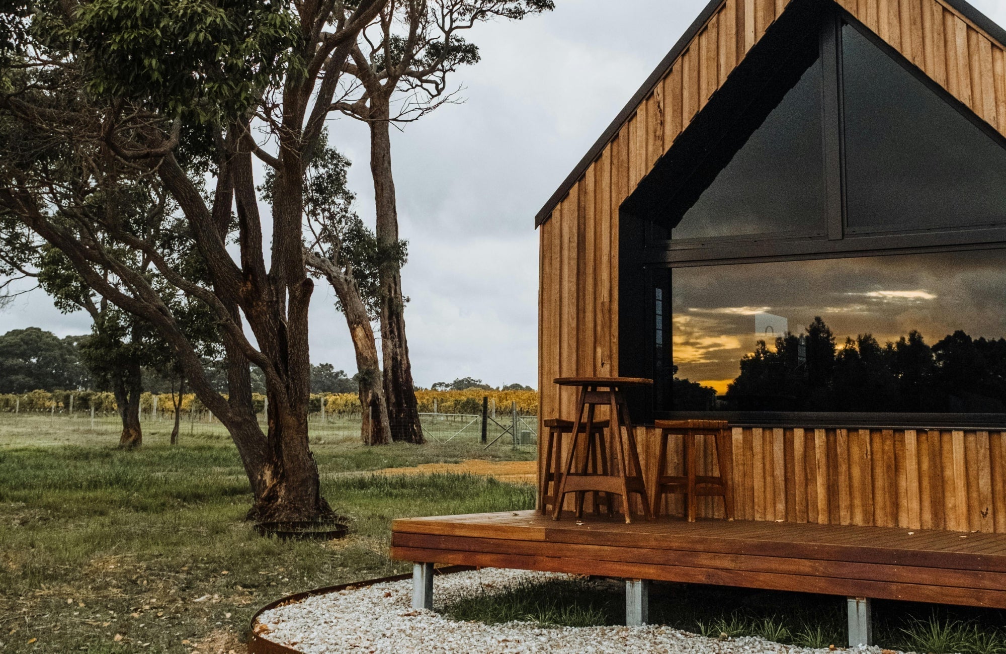 A modern wooden cabin with a large reflective window and a small outdoor deck featuring wooden counter stools overlooking a grassy landscape with tall trees at sunset.