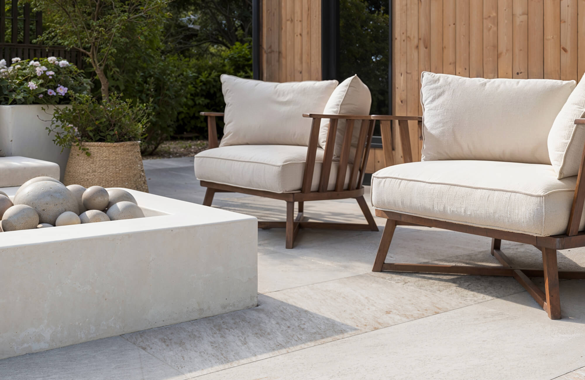 Modern outdoor patio furniture with two cushioned wooden chairs beside a minimalist concrete fire pit on a stone patio.