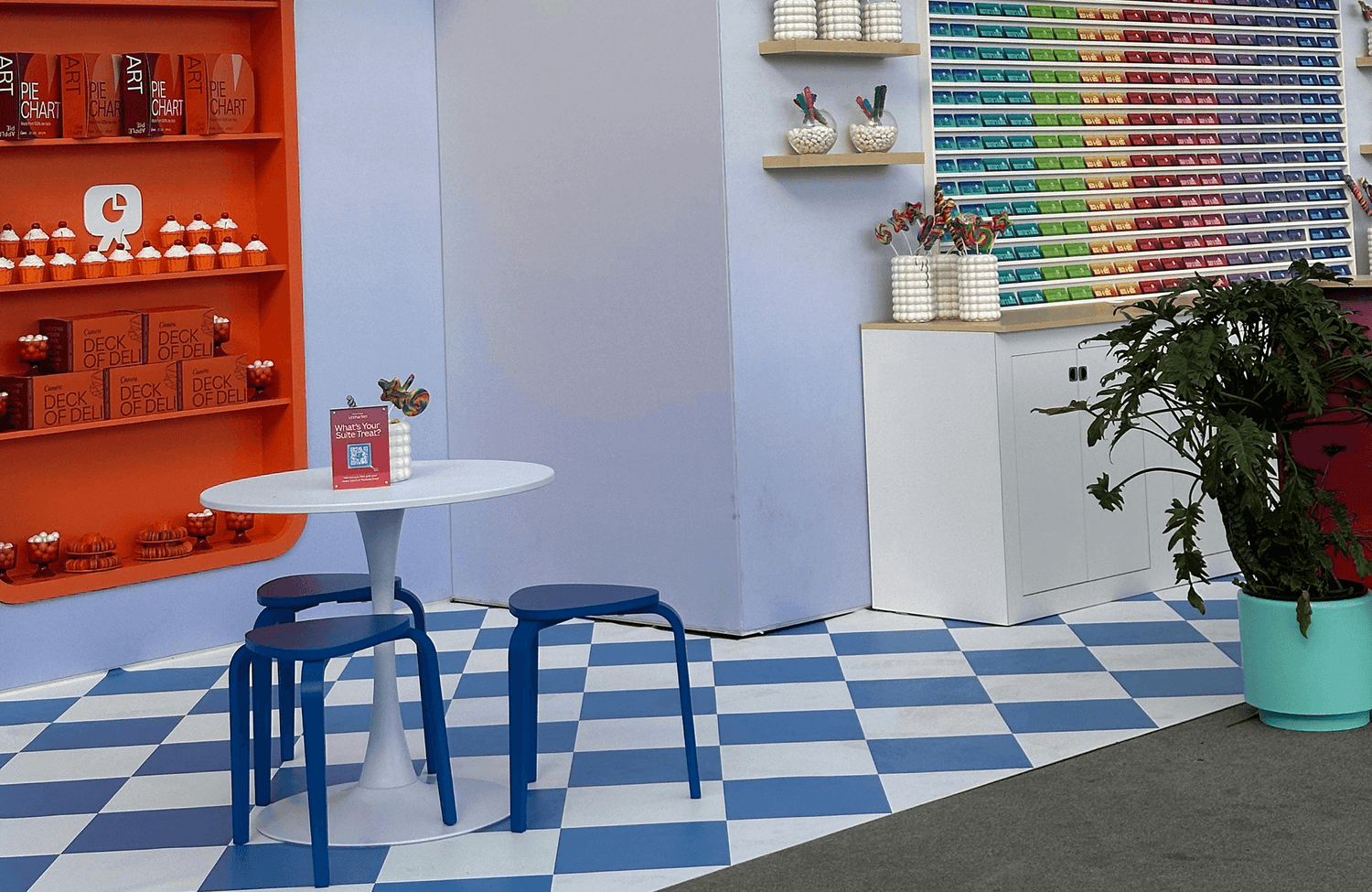 Playful blue and white checkerboard floor tiles beneath a vibrant candy-colored café setup with bold accents and colorful shelving.