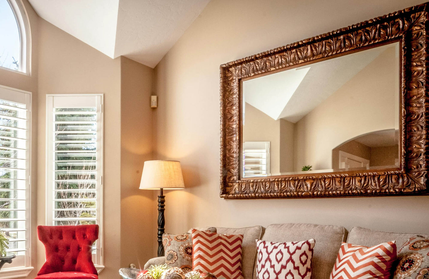 A large wall mirror with an ornate carved wood frame anchors the living room wall, reflecting warm natural light and complementing the neutral furnishings, patterned pillows, and inviting, traditional décor.