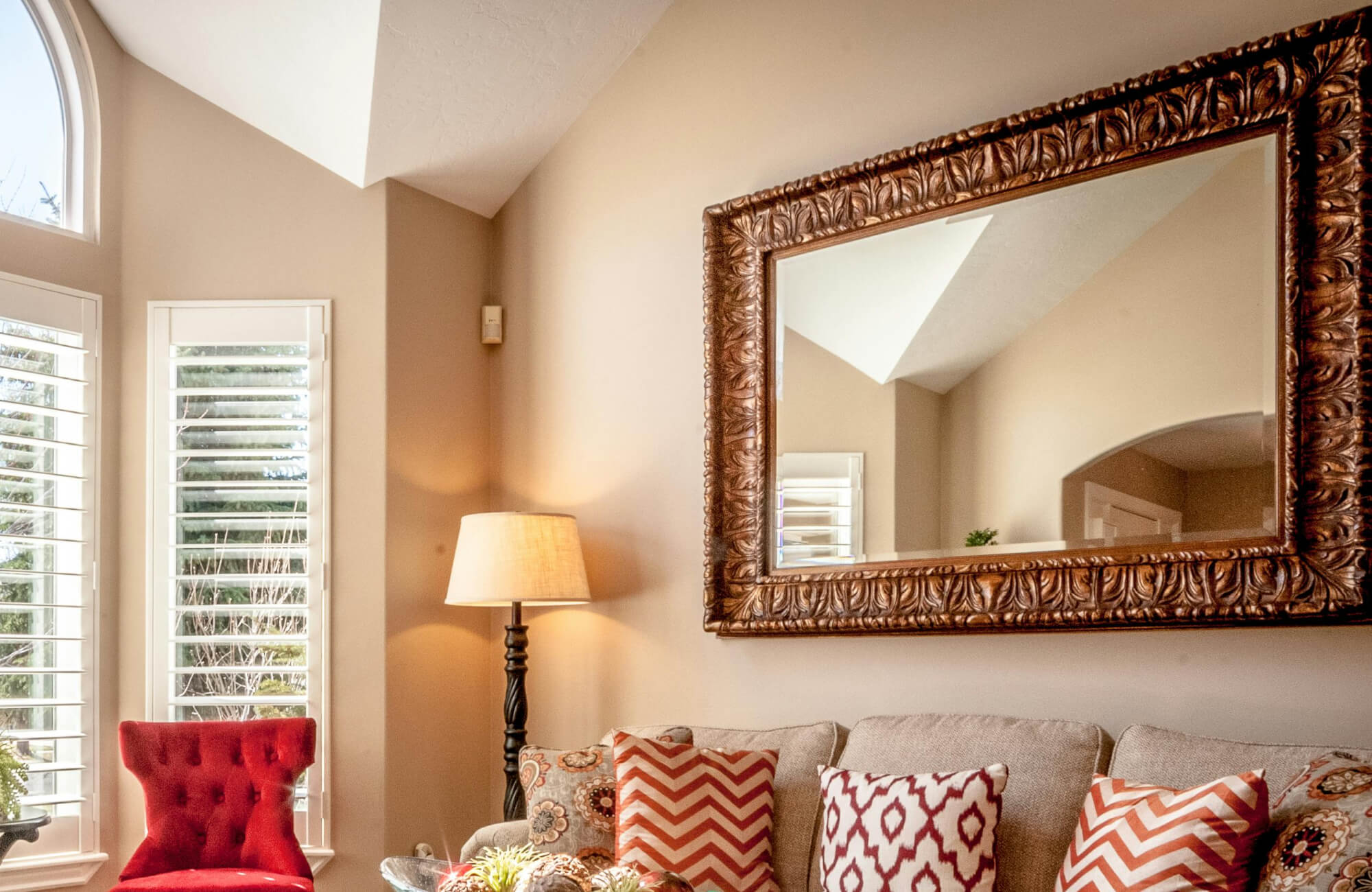 A large wall mirror with an ornate carved wood frame anchors the living room wall, reflecting warm natural light and complementing the neutral furnishings, patterned pillows, and inviting, traditional décor.