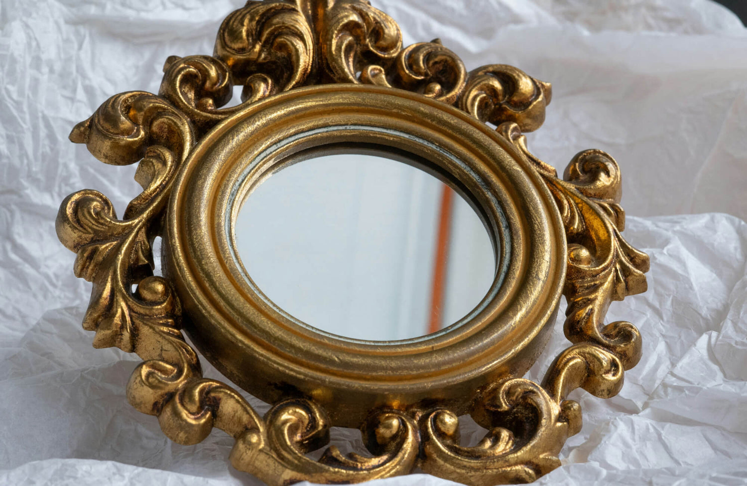Ornate round mirror with a gilded baroque-style frame featuring elaborate scrollwork on crumpled white paper.