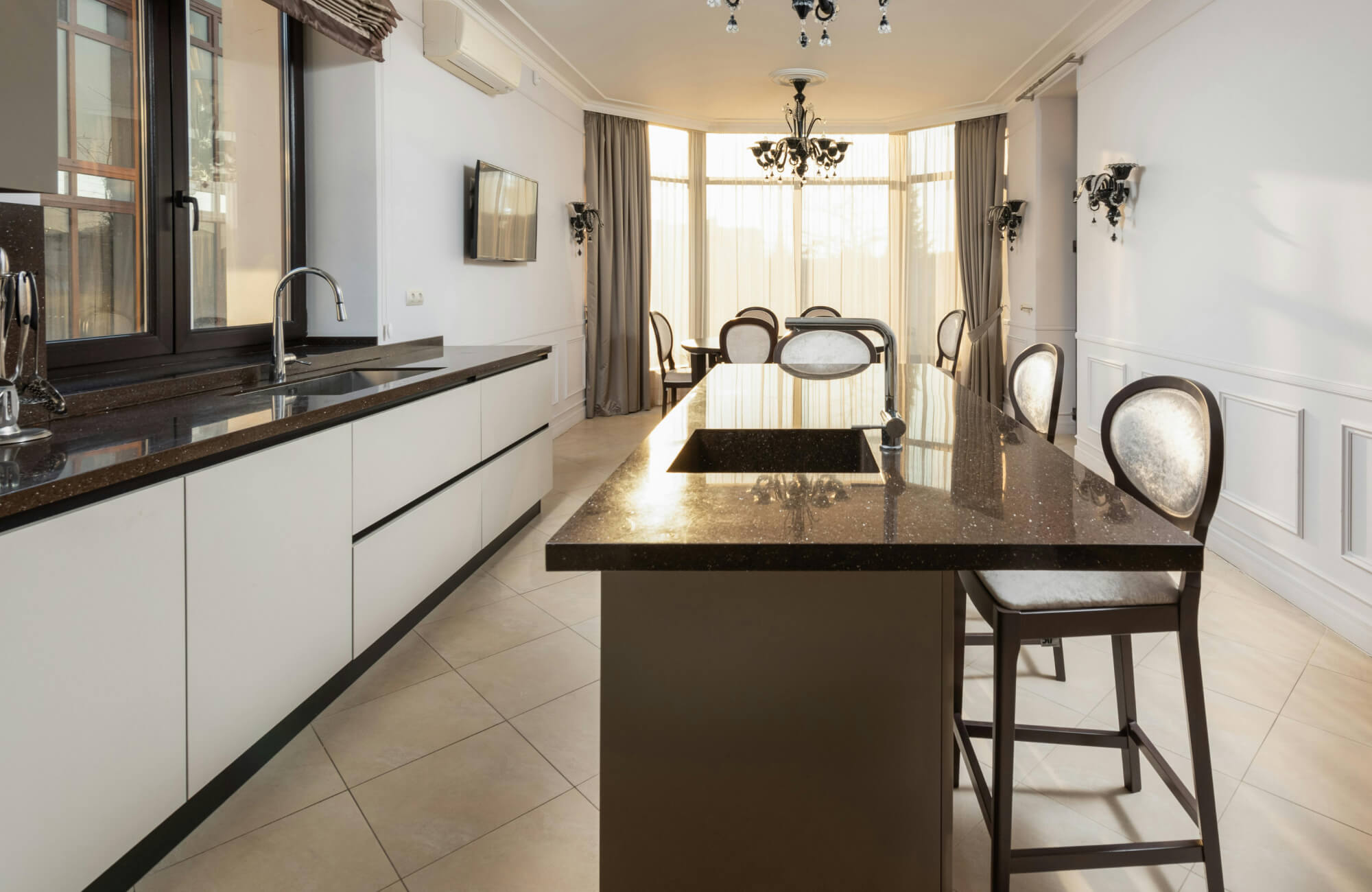 Bright, sophisticated open-concept kitchen and dining space featuring a sleek black granite island paired with a counter height dining set of upholstered chairs, complemented by white cabinetry and elegant chandeliers.