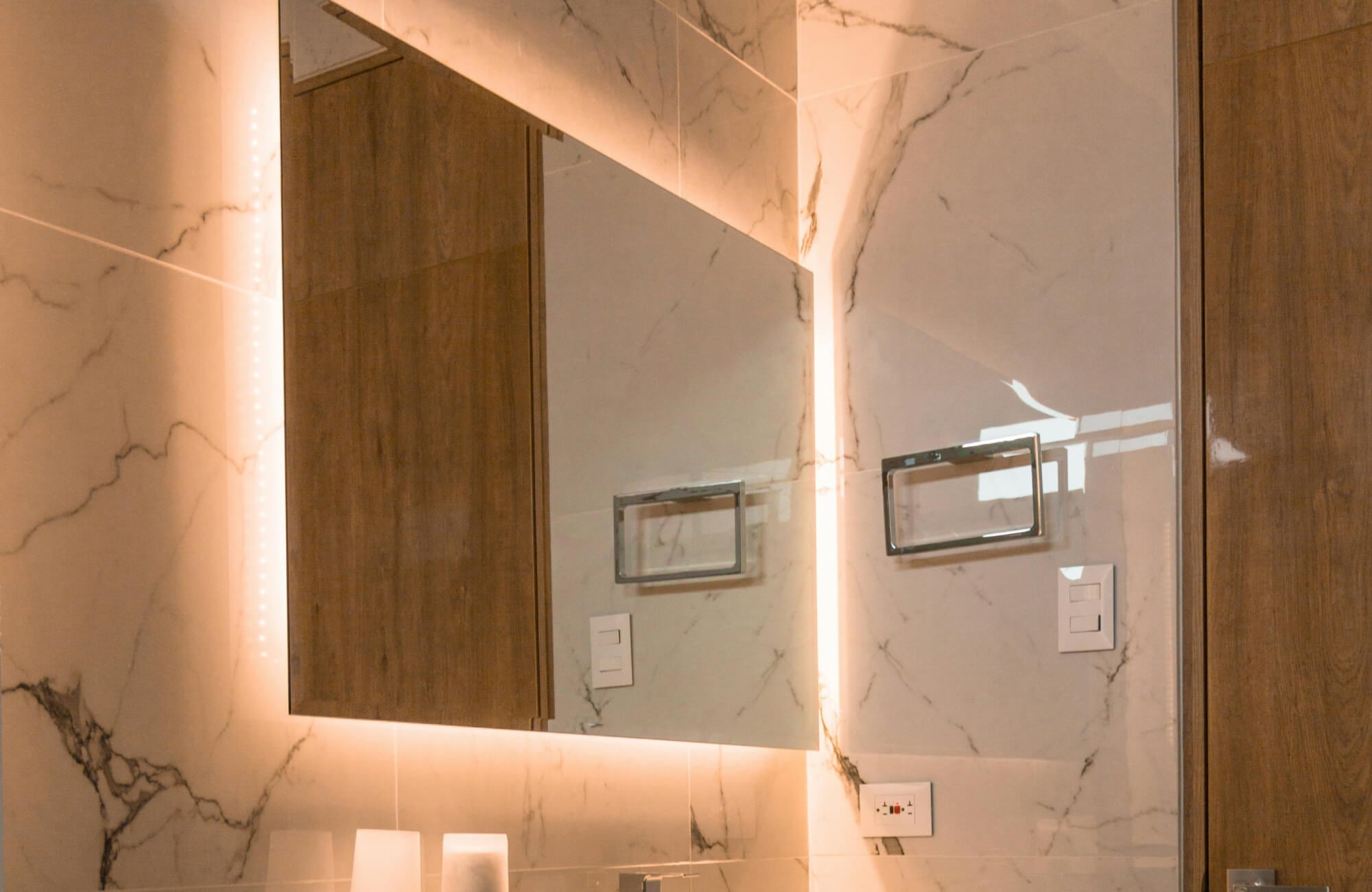 Frameless LED bathroom mirror with warm backlighting mounted on marble-look wall tiles, creating a soft glow that highlights the vanity area and surrounding finishes.