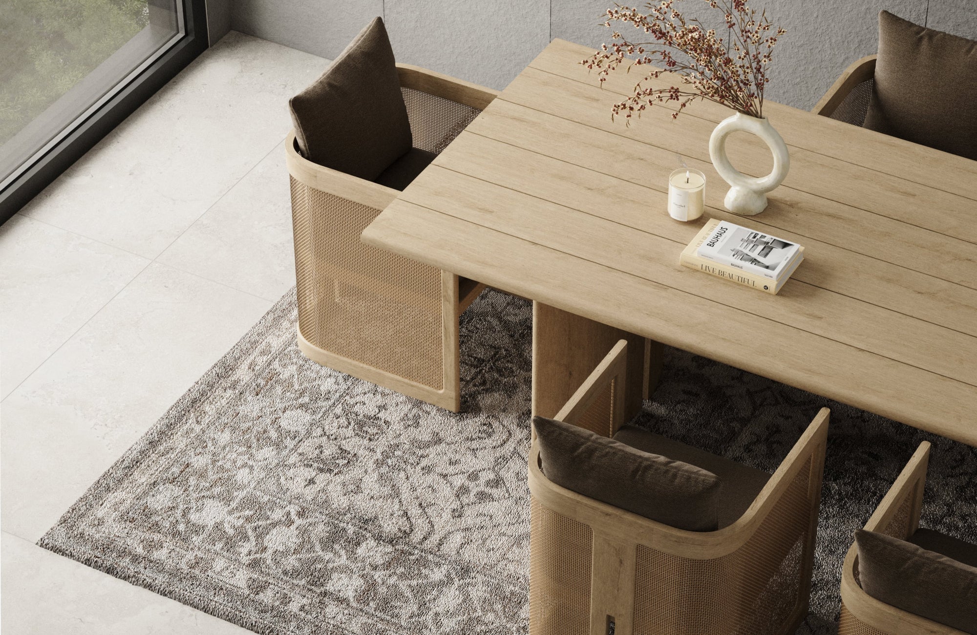 Dining area with light wood table and woven chairs set over a textured gray rug on pale stone flooring beside a window.