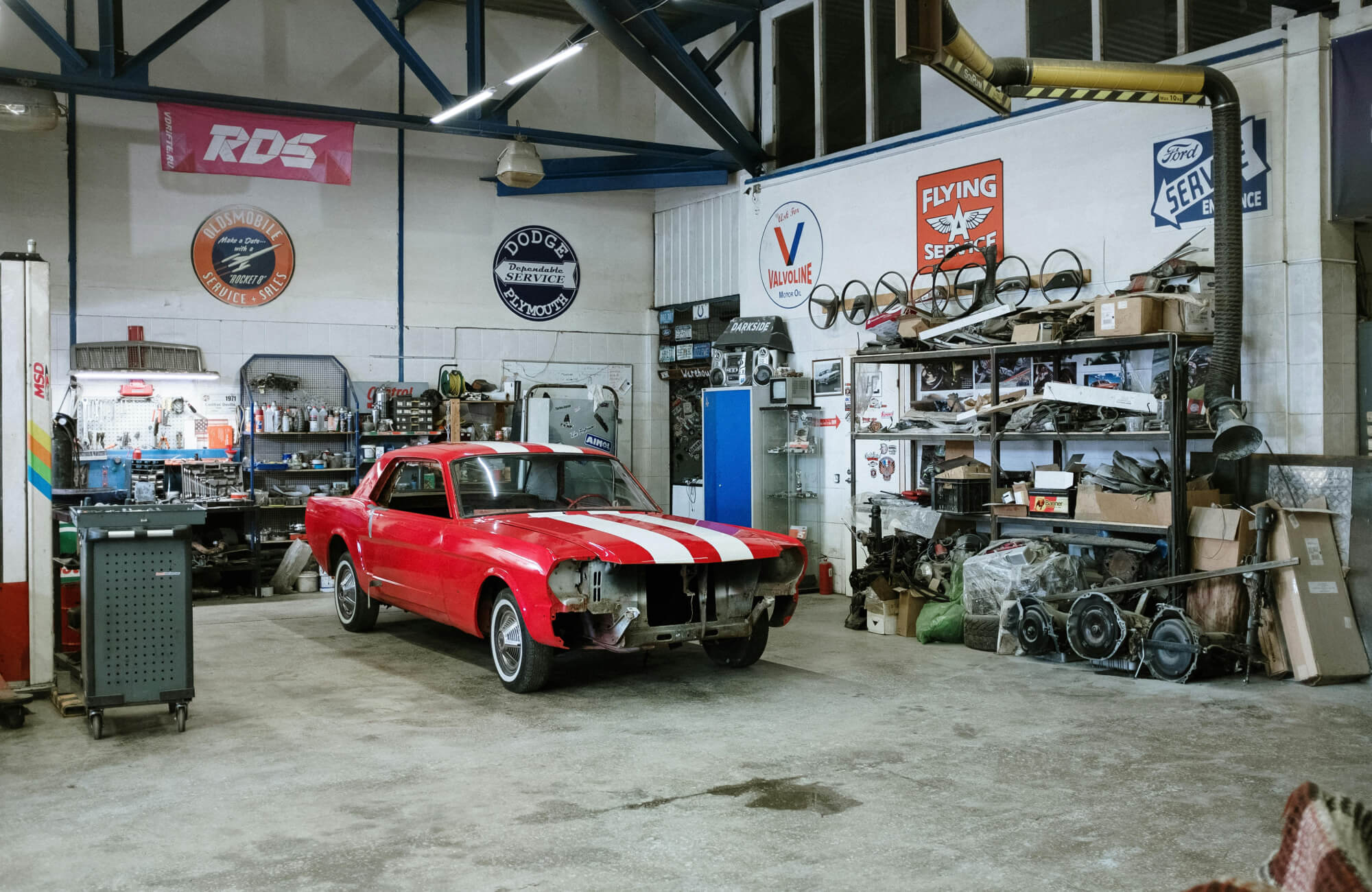 Red vintage car under restoration in a spacious automotive workshop, surrounded by tools, car parts, and classic service signs on the walls.