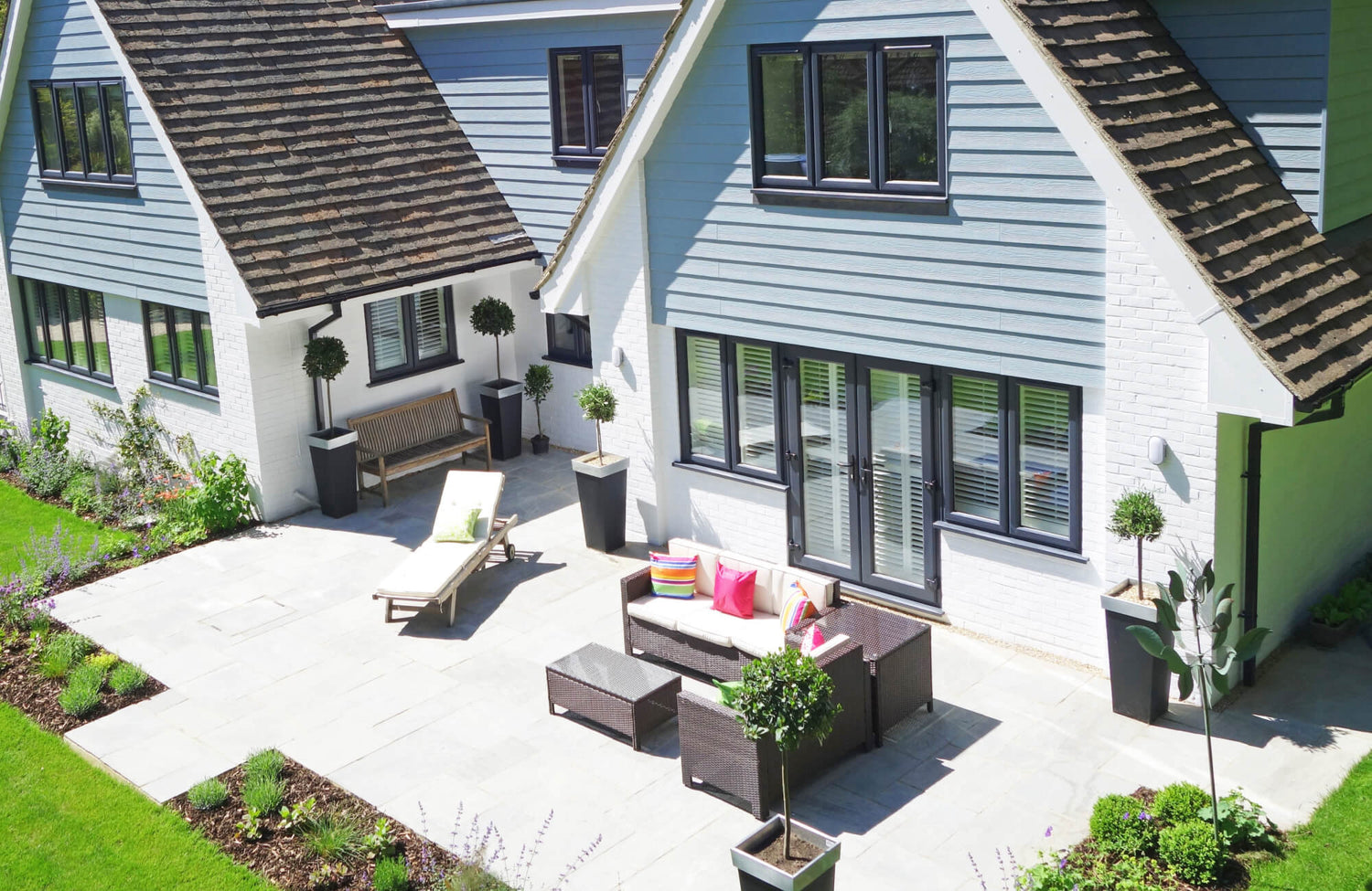 A bright, modern patio features expansive light-gray outdoor tile flooring that anchors wicker seating, potted greenery, and a sun lounger against the home’s crisp white brick and blue siding exterior.