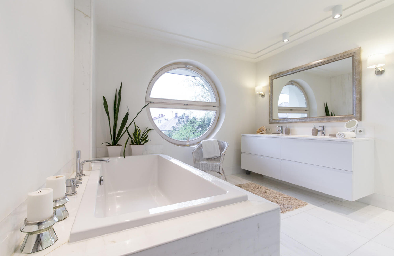 Bright contemporary bathroom with a round window, wall sconces beside a large mirror, and a white tub accented by green plants.
