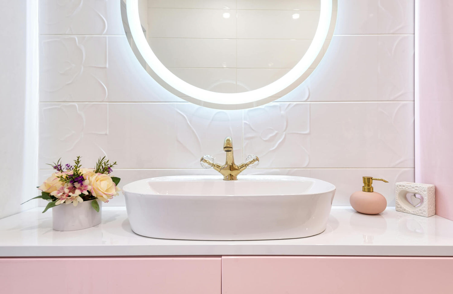 Soft backlit round mirror glowing above a pink vanity and floral tile wall in a bright bathroom.