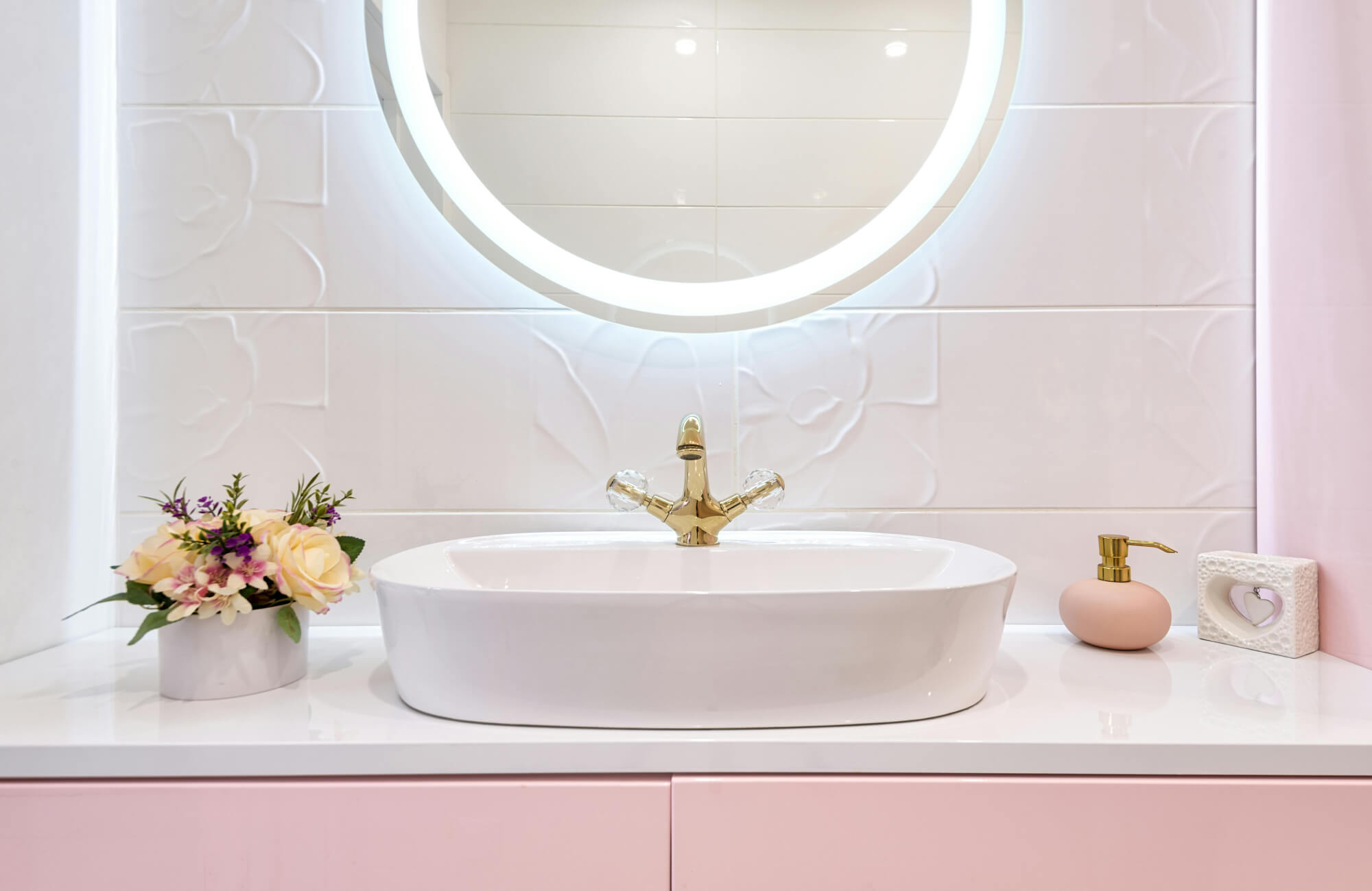 Soft backlit round mirror glowing above a pink vanity and floral tile wall in a bright bathroom.