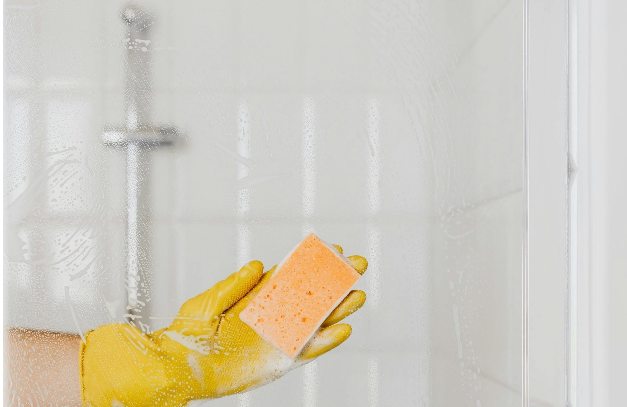 A close-up view shows someone cleaning a glass shower door with an orange sponge, revealing white shower tile blurred in the background.