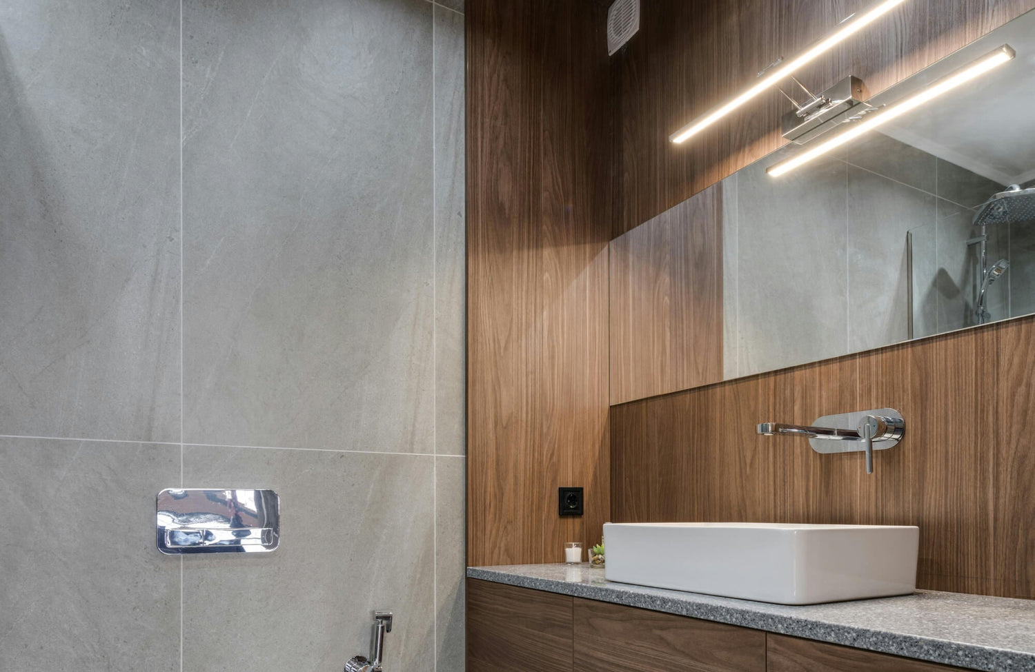 A modern bathroom vanity scene featuring a sleek horizontal vanity light above a large frameless mirror, paired with warm wood cabinetry and large-format gray stone-look wall tile.