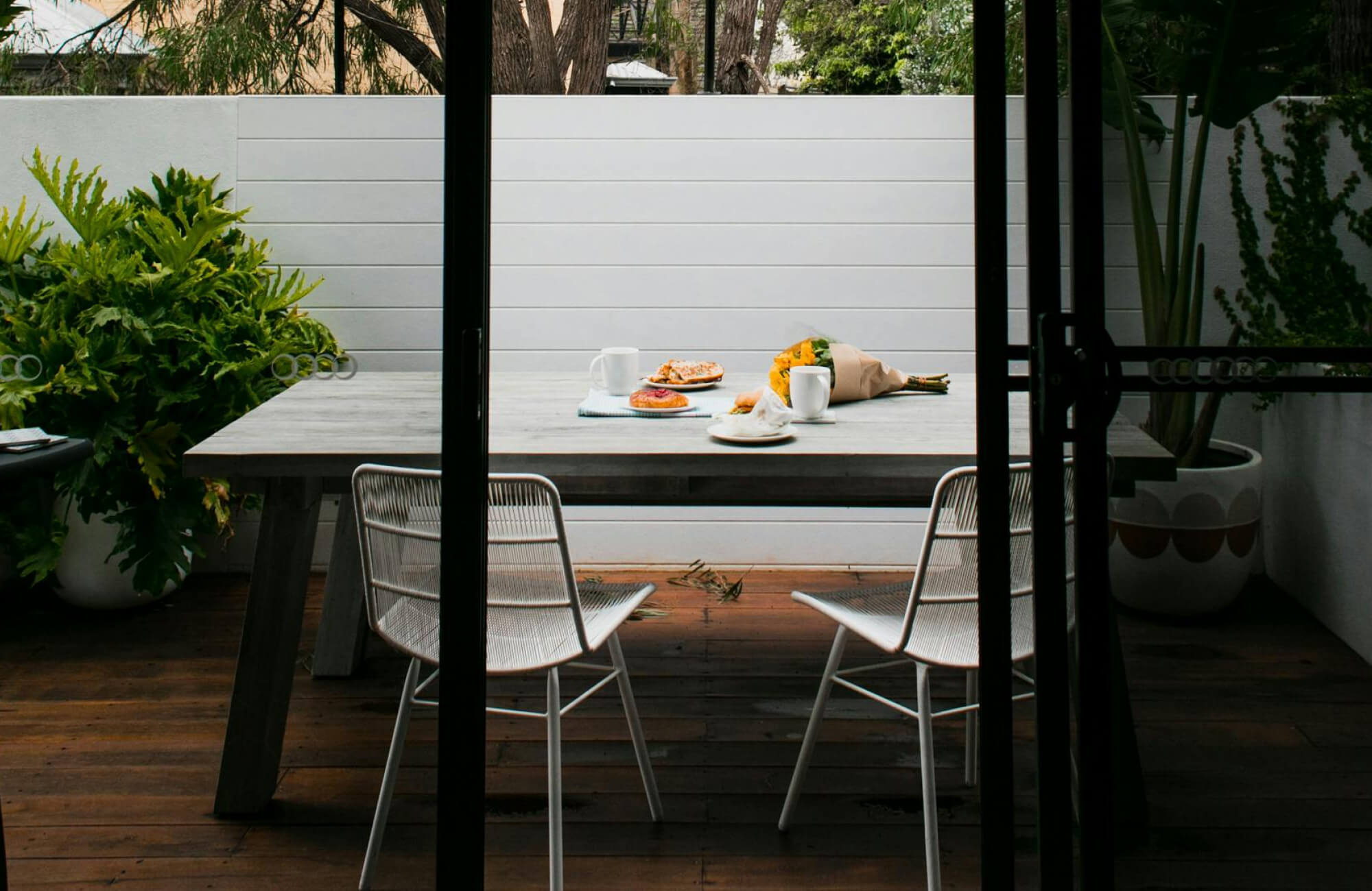 Cozy outdoor dining area with a rustic wood table, white chairs, fresh pastries, and a bouquet of sunflowers.
