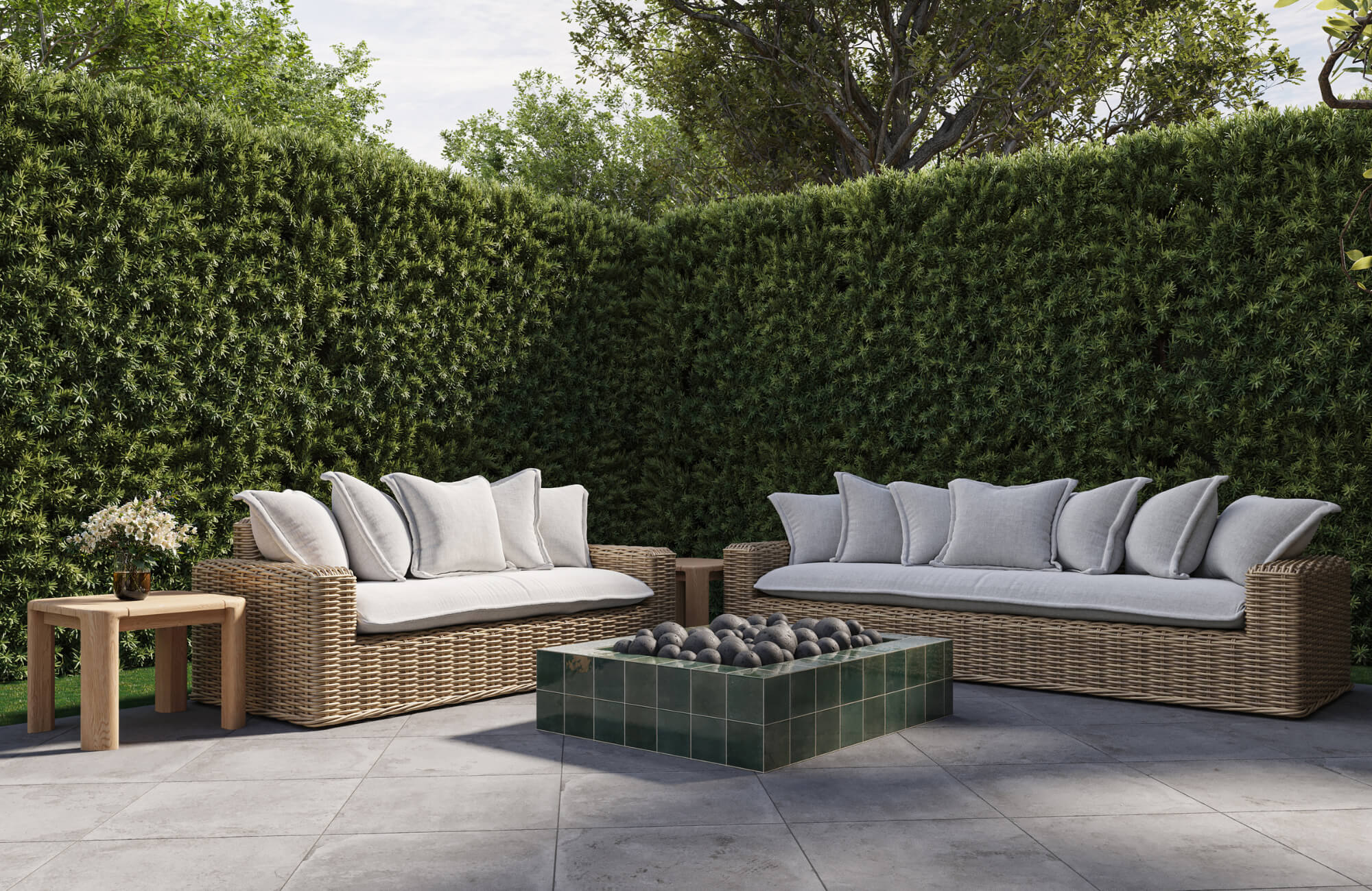 Outdoor wicker sofas styled with neutral pillows around a tiled fire pit, set against lush green hedges for a refined lounge setting.