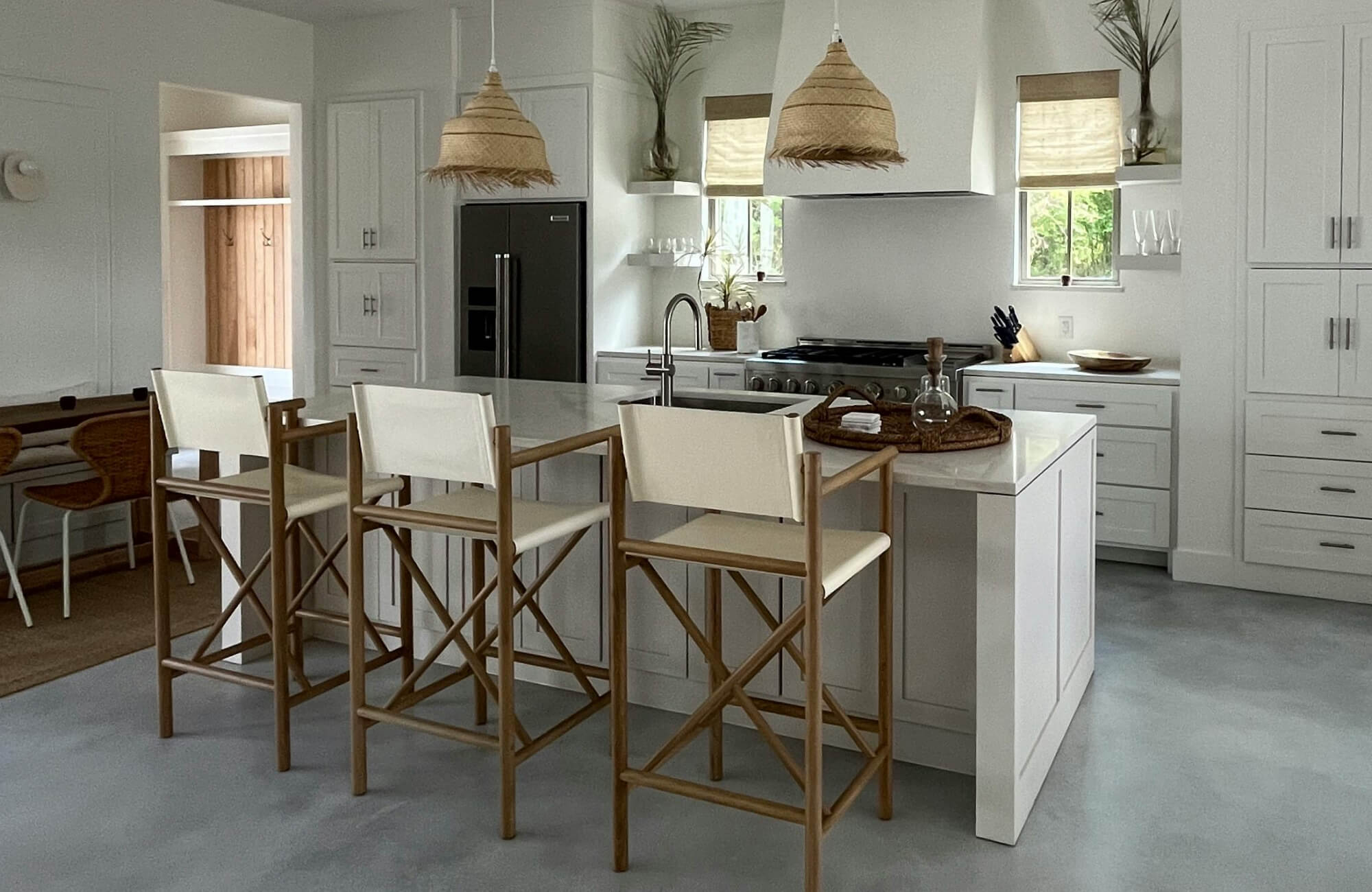 A serene, coastal-inspired kitchen showcases white cabinetry, natural wood seating, and a spacious island illuminated by two woven kitchen pendant lights that bring warmth, texture, and relaxed elegance to the space.