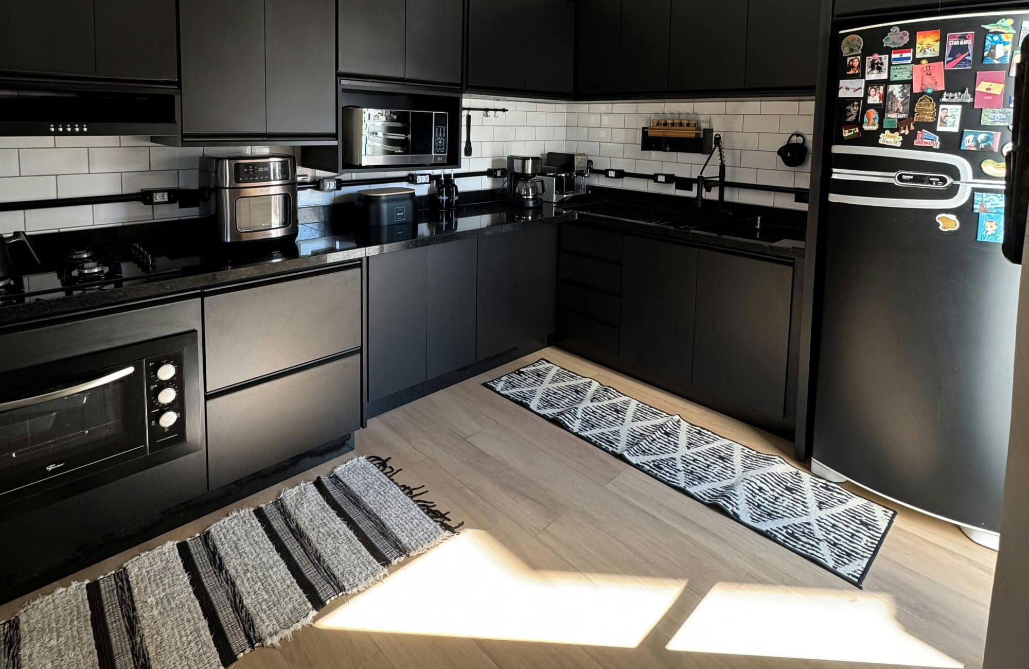 A modern black kitchen is accented by a geometric kitchen rug runner, adding visual softness and comfort along light wood flooring while contrasting sleek matte cabinetry, white subway tile backsplash, and integrated appliances.
