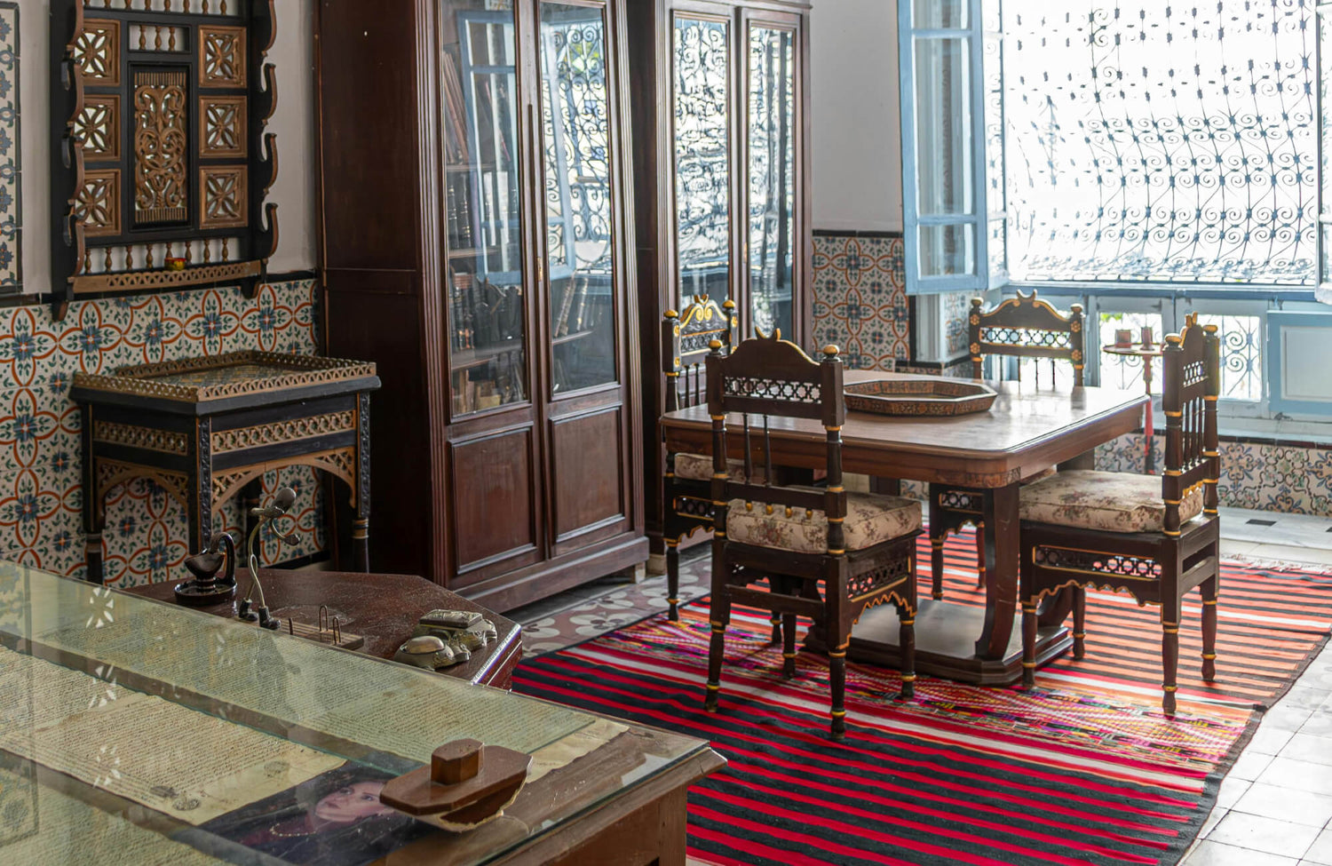 Traditional Moroccan-style room with patterned tile walls, carved wood furniture, and a striking red patterned rug.