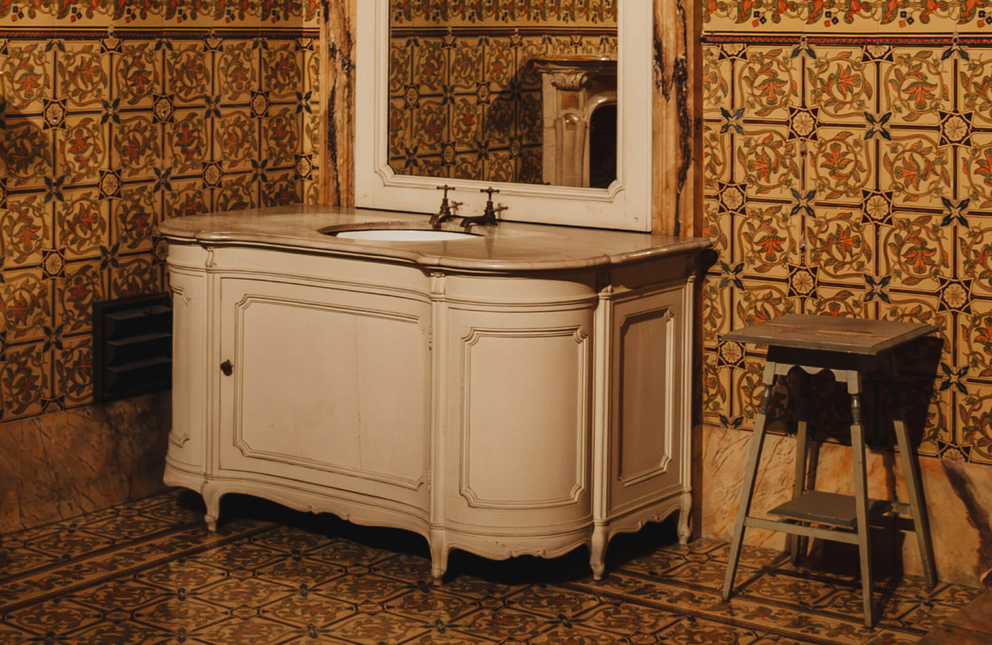 Vintage bathroom with ornate floral-patterned tiles on walls and floor, paired with an antique cream vanity and mirror.