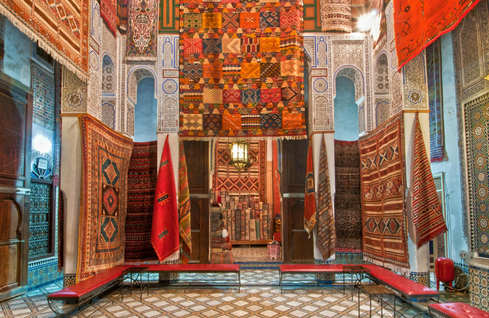 Traditional Moroccan rug shop with vibrant handwoven carpets adorning intricate mosaic walls and patterned tile flooring.