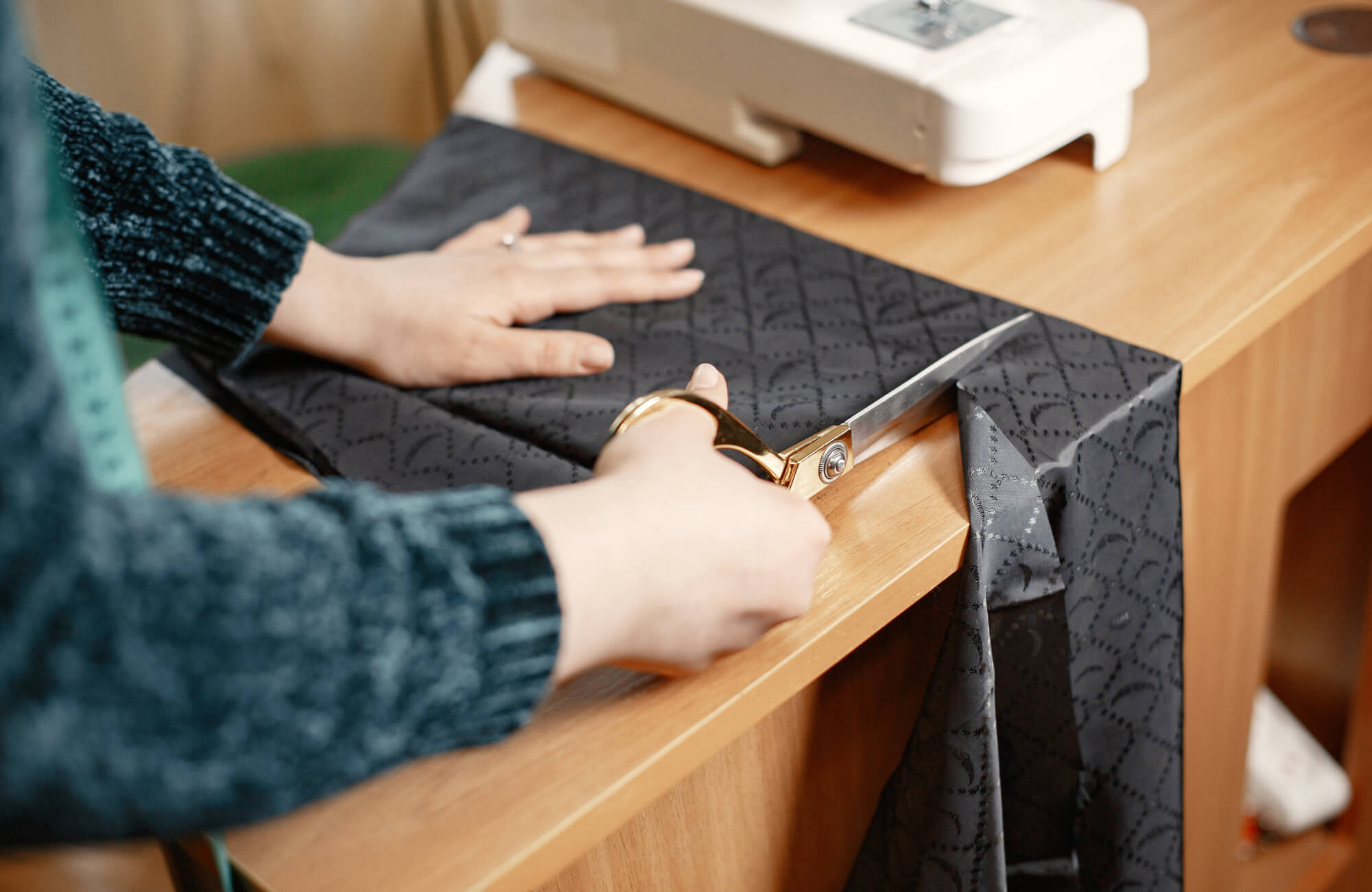 Cutting patterned fabric with large tailoring scissors on a wooden work table, preparing textile pieces for sewing decorative pillows or home décor accents.