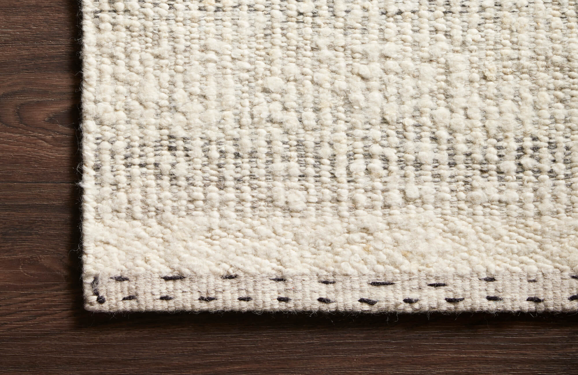 Detailed view of a neutral wool-blend rug showing looped texture, subtle gray patterning, and stitched edge against a dark hardwood floor.