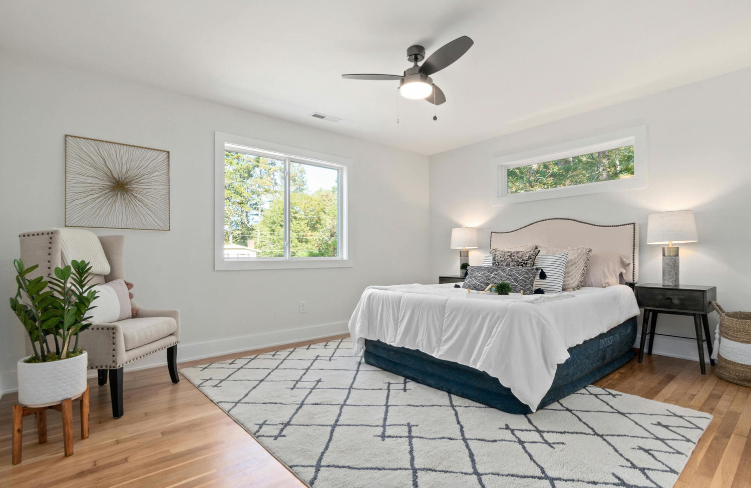 Modern bedroom featuring a king bed layered with pillows, a textured white rug, black nightstands with lamps, and sunlight streaming through two large windows.