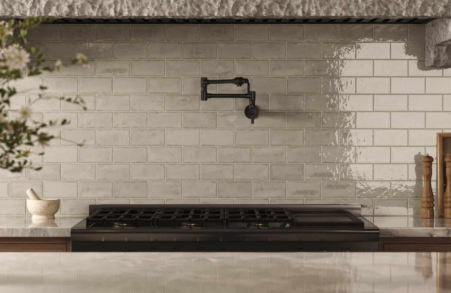 A kitchen backsplash with glossy ceramic subway tiles in soft gray tones, paired with a black pot filler above a gas range and light marble countertops.
