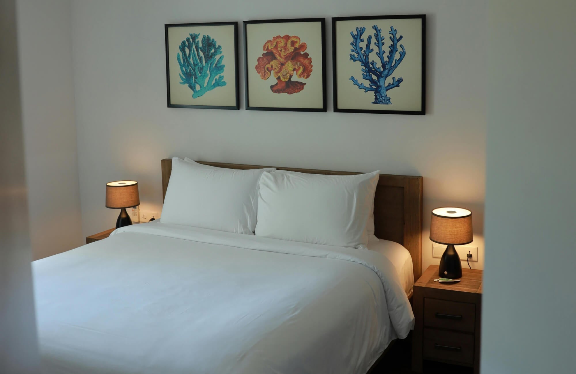 Minimalist bedroom with crisp white bedding, warm bedside lighting, and framed coral-inspired wall art in calming blue and orange tones above a wooden headboard.