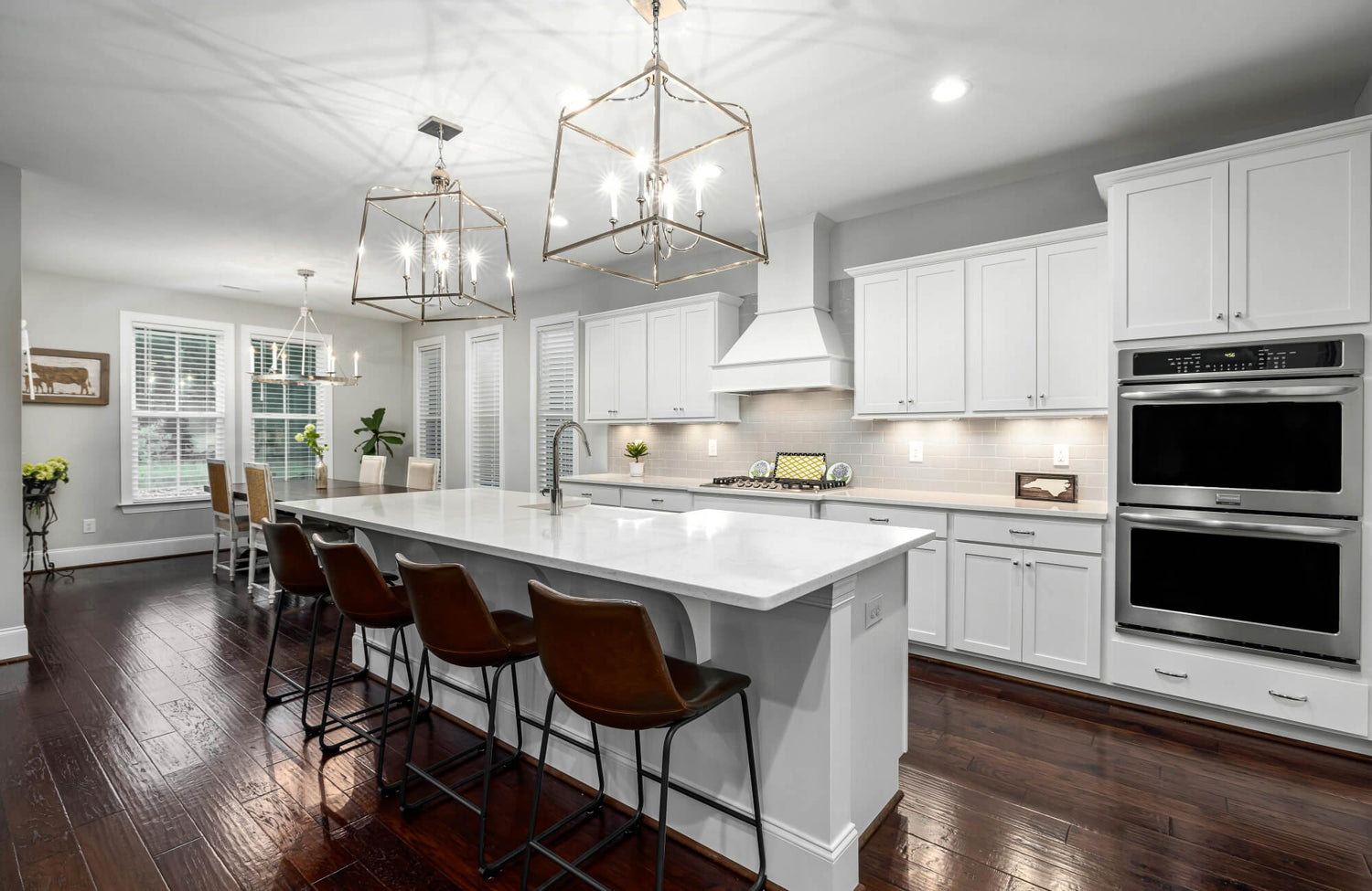 Bright modern kitchen with a large white island, backless bar stools, and geometric pendant lights, surrounded by shaker cabinets and warm wood flooring.