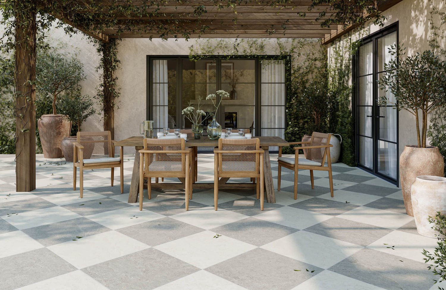 Porcelain paver terrace in a neutral checker pattern, styled with woven wood chairs, rustic dining table, and Mediterranean-inspired foliage.