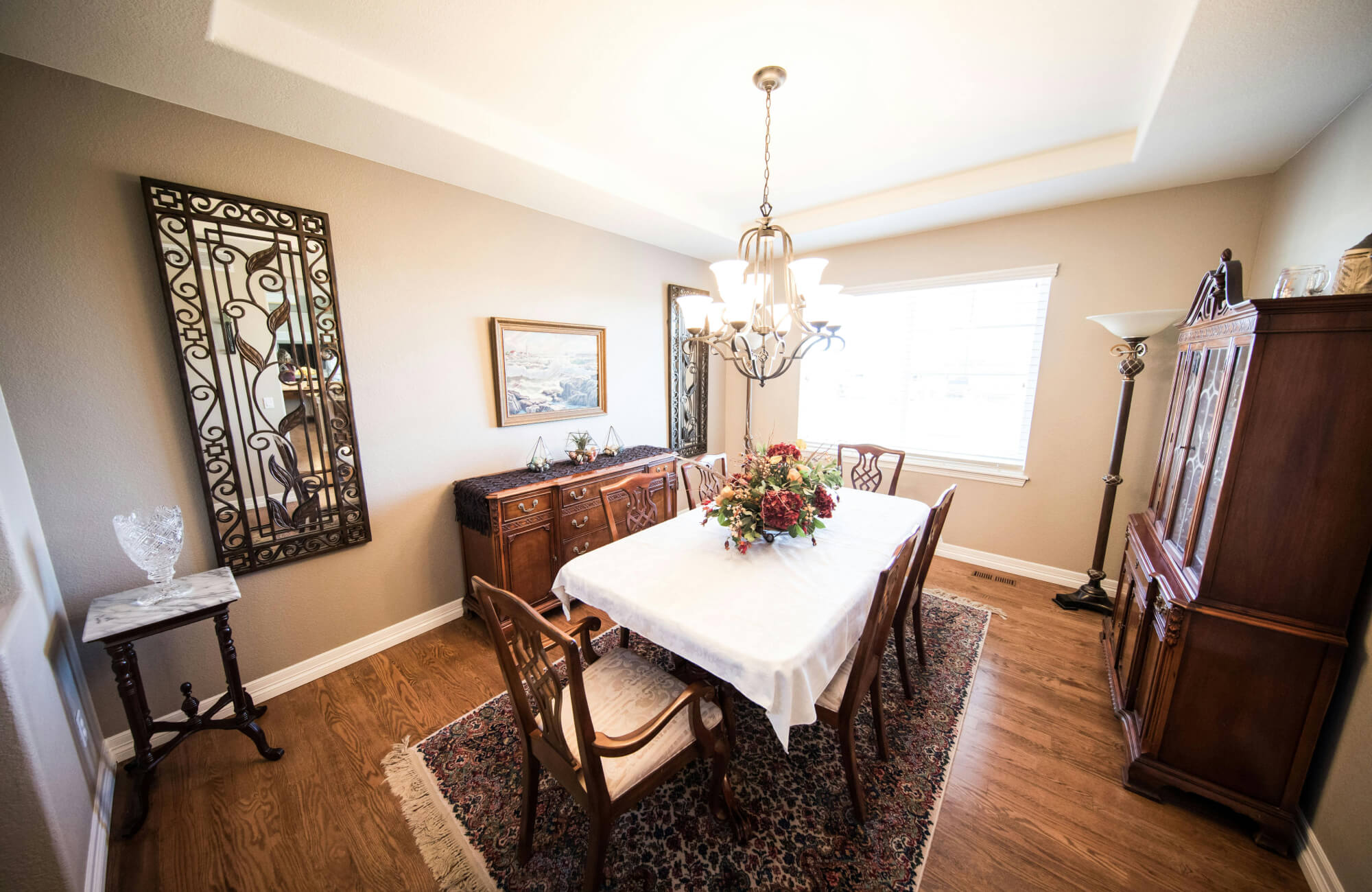 Traditional dining room with a six-seat wooden table on a patterned area rug, surrounded by classic wood furniture and a chandelier overhead.