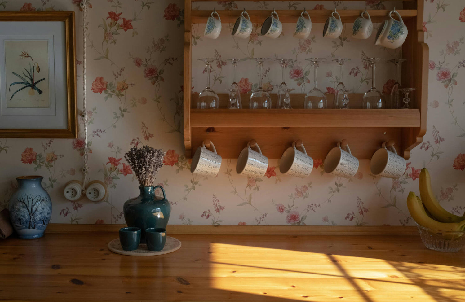 Cozy kitchen corner with floral wallpaper, wooden shelf holding mugs and glassware, warm sunlight casting shadows across a wooden countertop.
