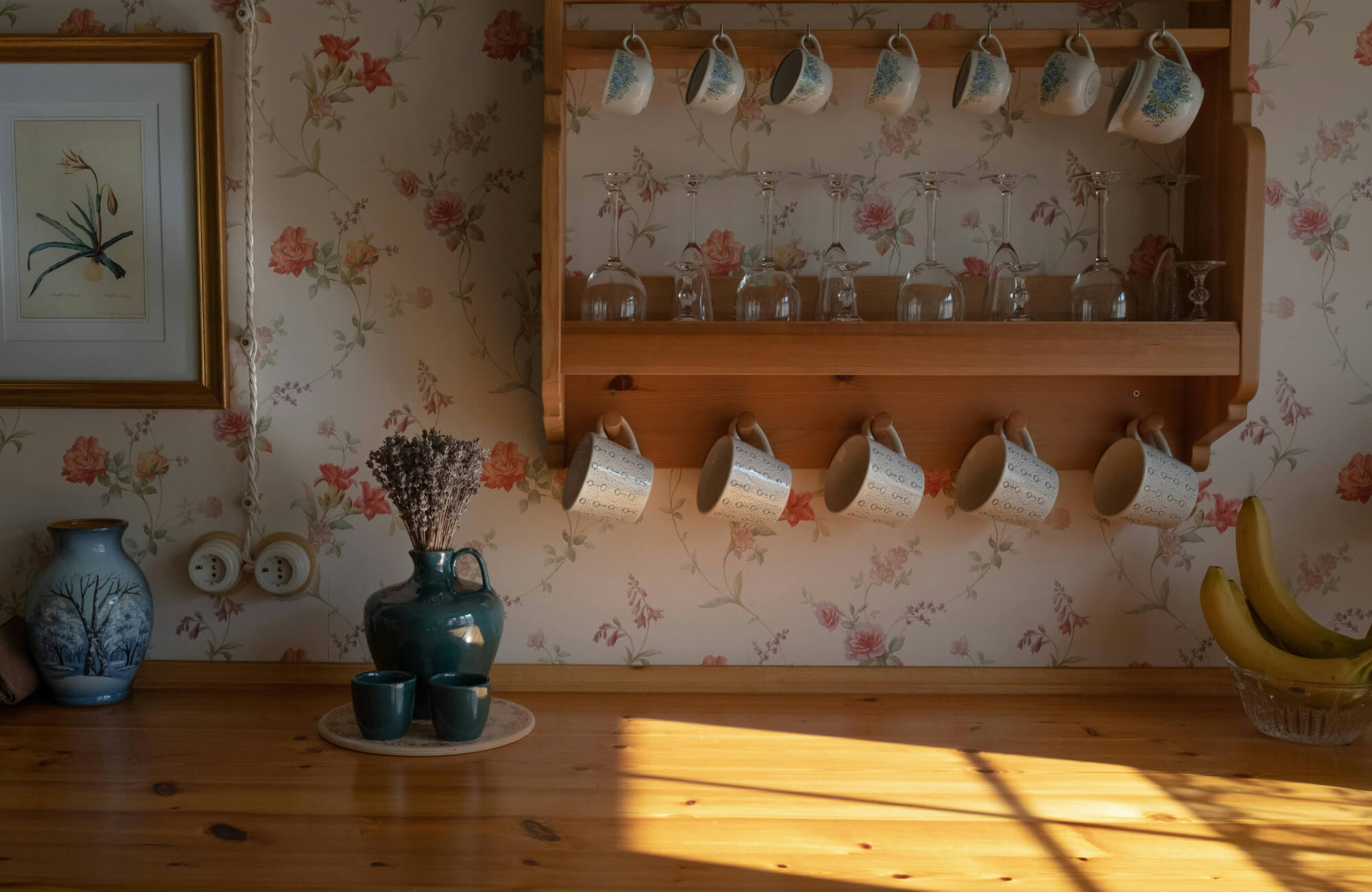 Cozy kitchen corner with floral wallpaper, wooden shelf holding mugs and glassware, warm sunlight casting shadows across a wooden countertop.