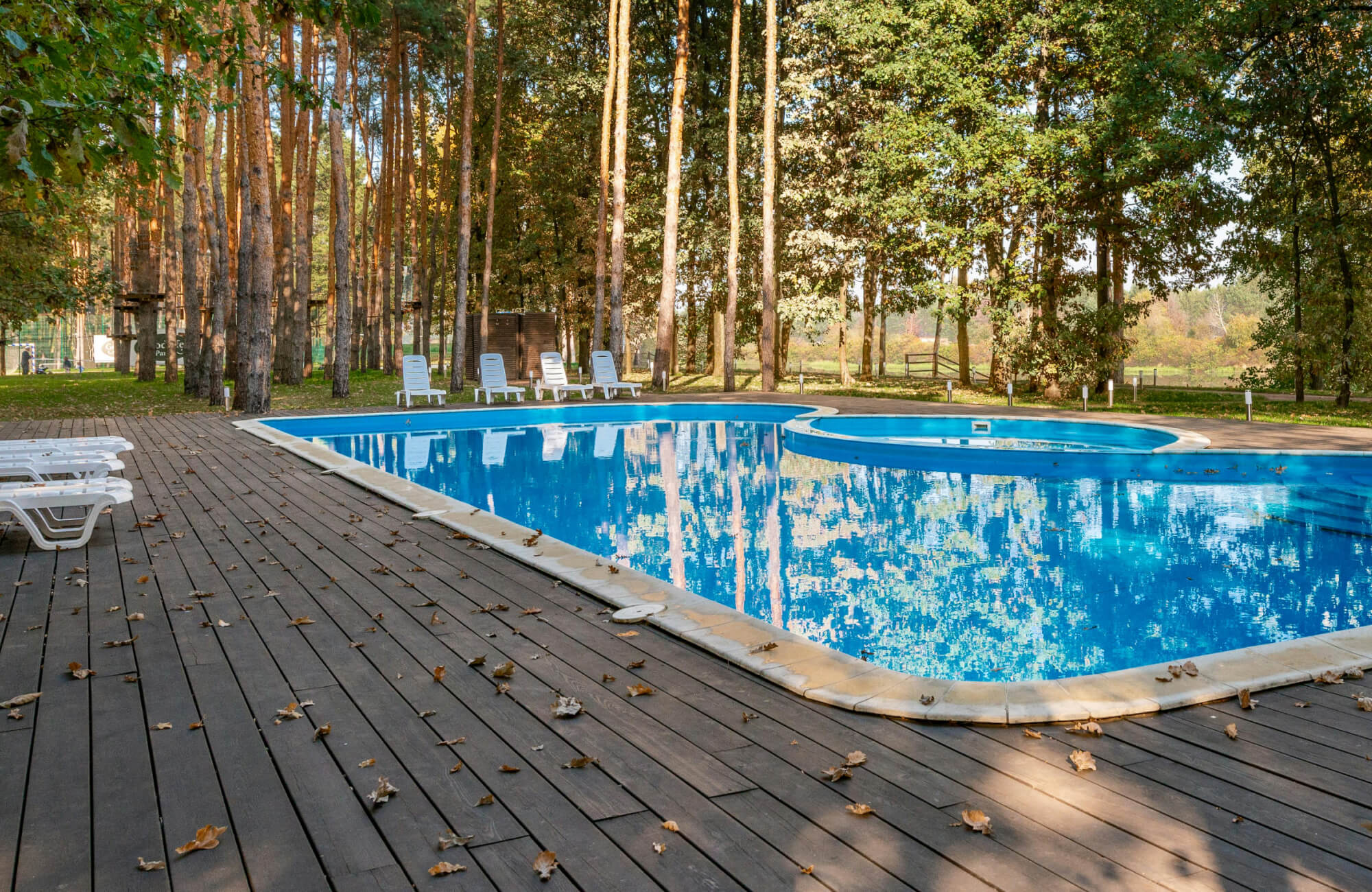 Hardwood deck flooring surrounding an outdoor swimming pool with lounge chairs, scattered leaves, and tall trees in the background.