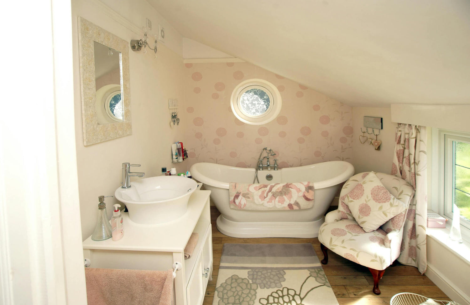 Cozy attic-style master bathroom with freestanding tub, compact vanity, soft pink wallpaper, and natural light from round window.