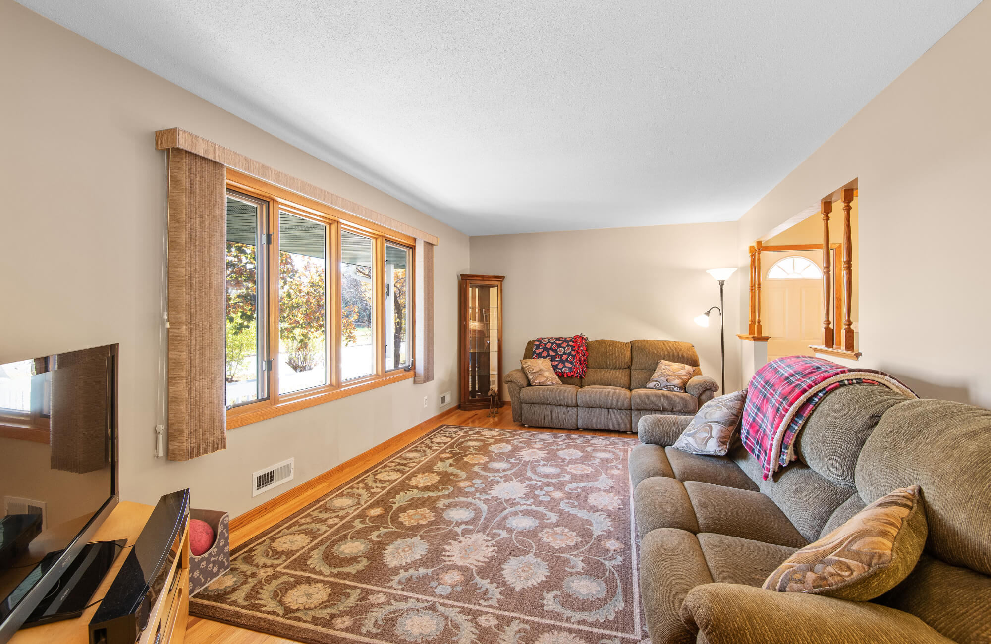 Warm-toned living room with large windows, neutral walls, brown floral area rug, and earth-toned upholstered sofas with throw pillows.