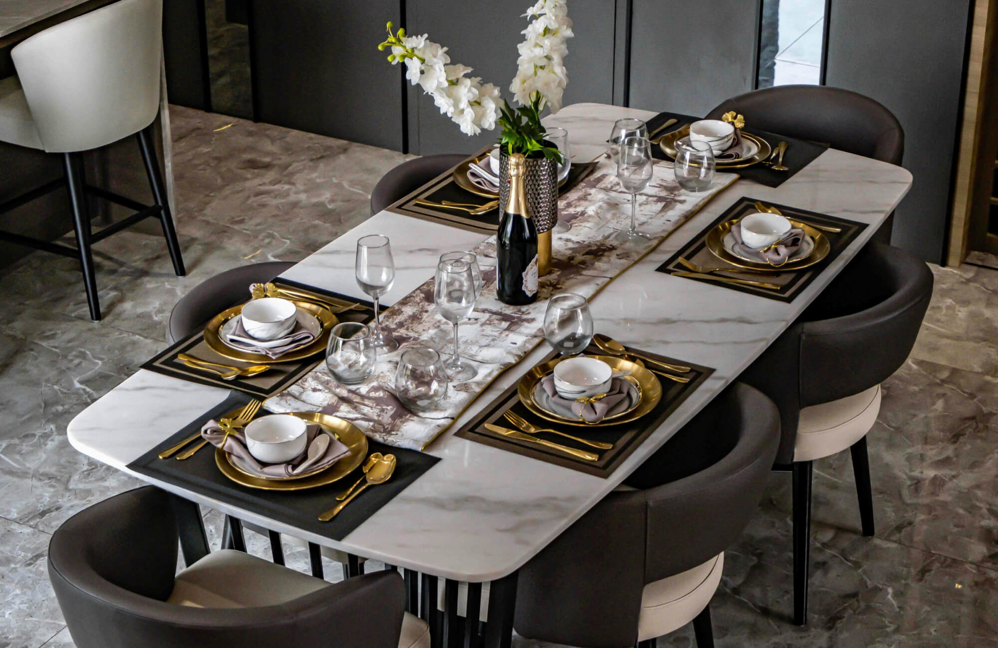 A contemporary marble dining table with subtle veining, styled with gold-accented place settings and dark upholstered dining chairs for a balanced, upscale feel.