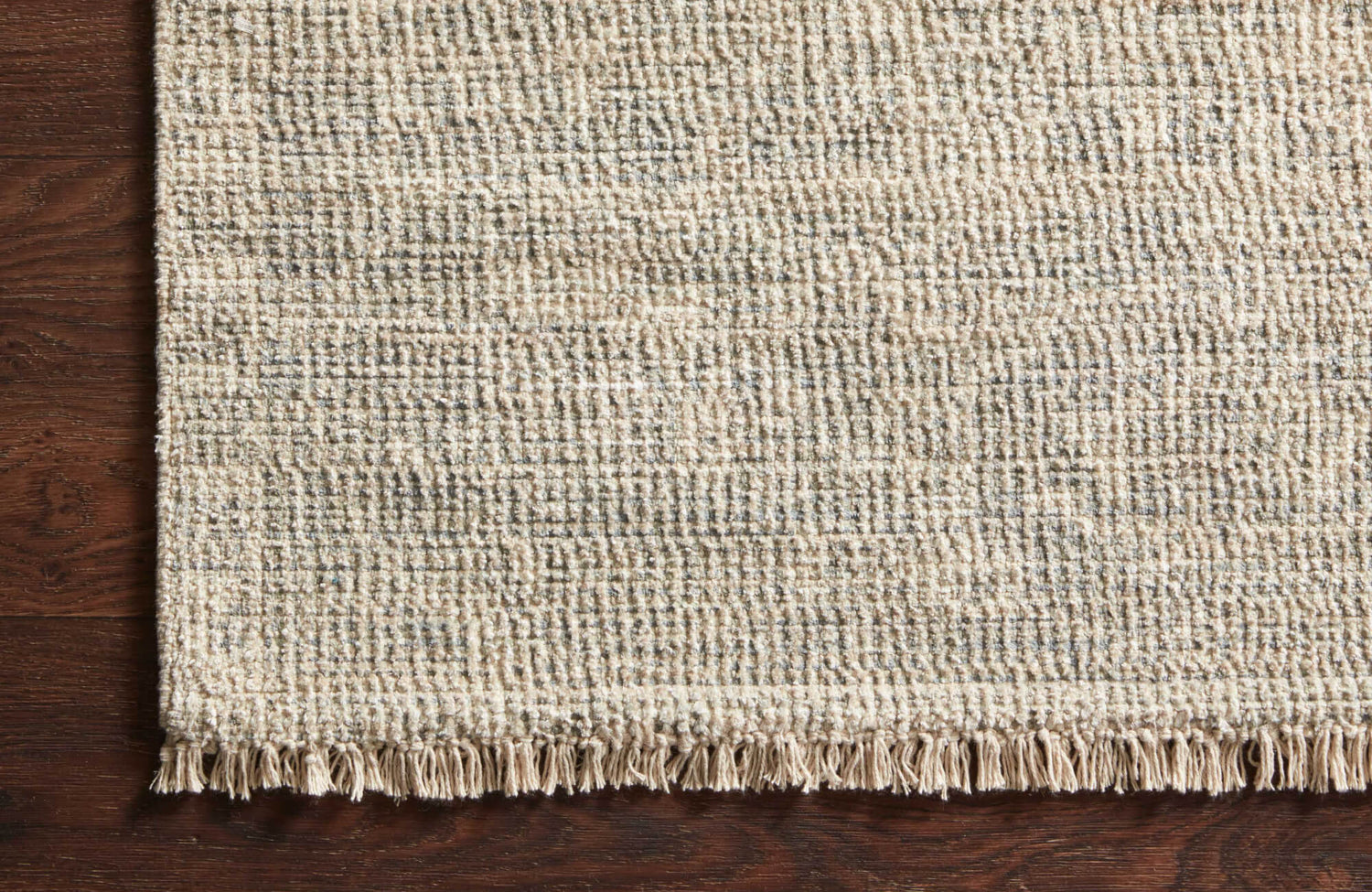 Textured flatweave rug in neutral tones with delicate fringe detailing, placed over a rich brown hardwood surface.
