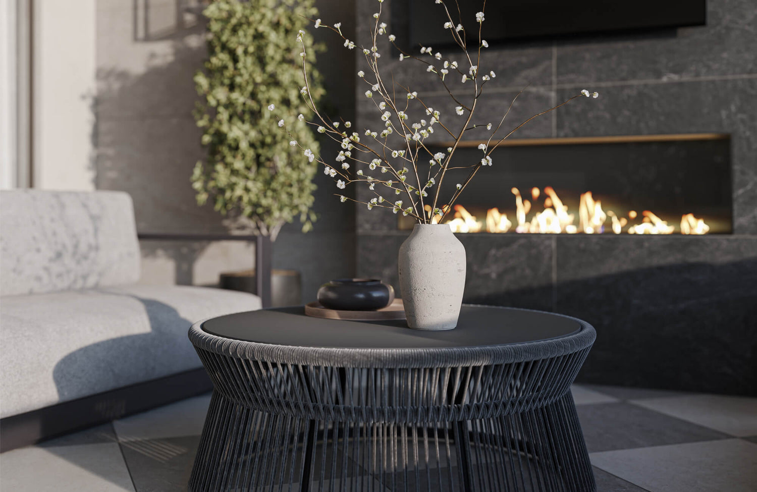 Modern outdoor lounge area with a woven round coffee table, light upholstered seating, floral vase, greenery, and a dark tiled fireplace wall with a linear fire feature.