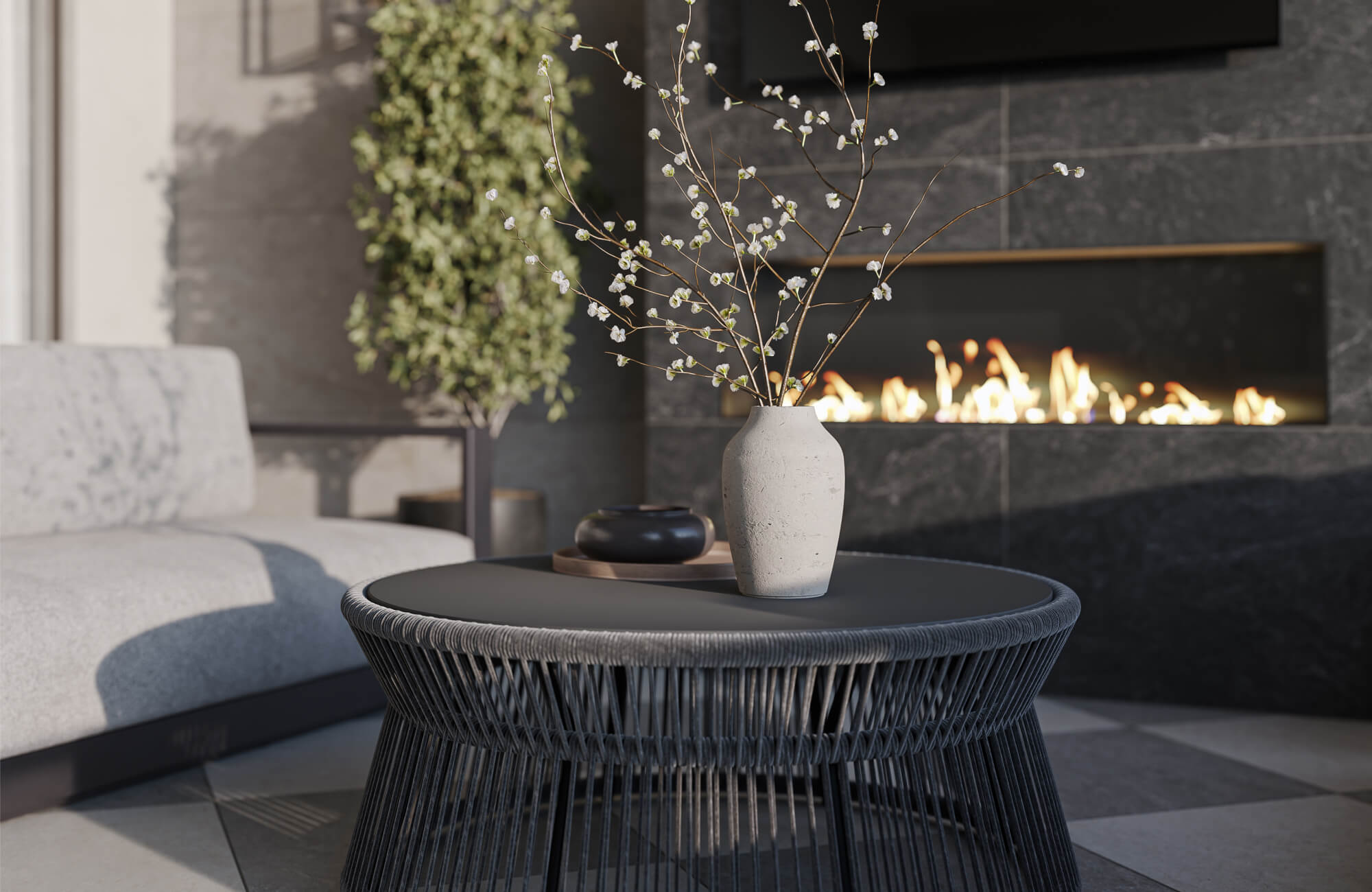 Modern outdoor lounge area with a woven round coffee table, light upholstered seating, floral vase, greenery, and a dark tiled fireplace wall with a linear fire feature.