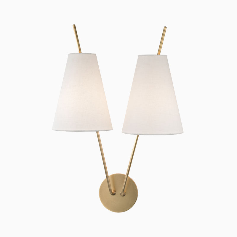 Aged brass wall sconce with round backplate, dual angled arms, and tapered white fabric shades for a modern balanced symmetrical design.
