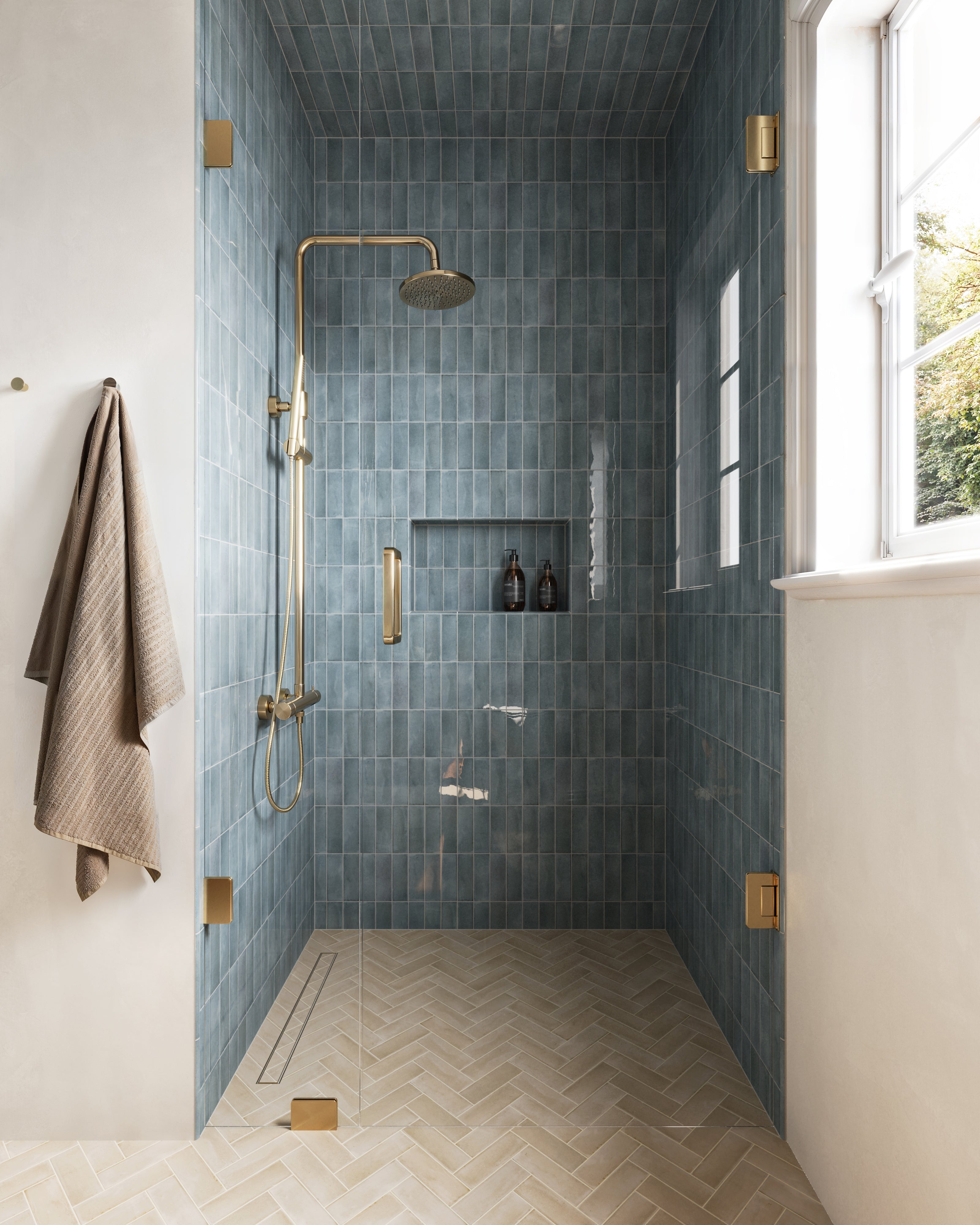 The shower features glossy blue tiles extending across the walls and ceiling, paired with brass fixtures and herringbone-patterned beige floor tiles for a stylish and luxurious look.