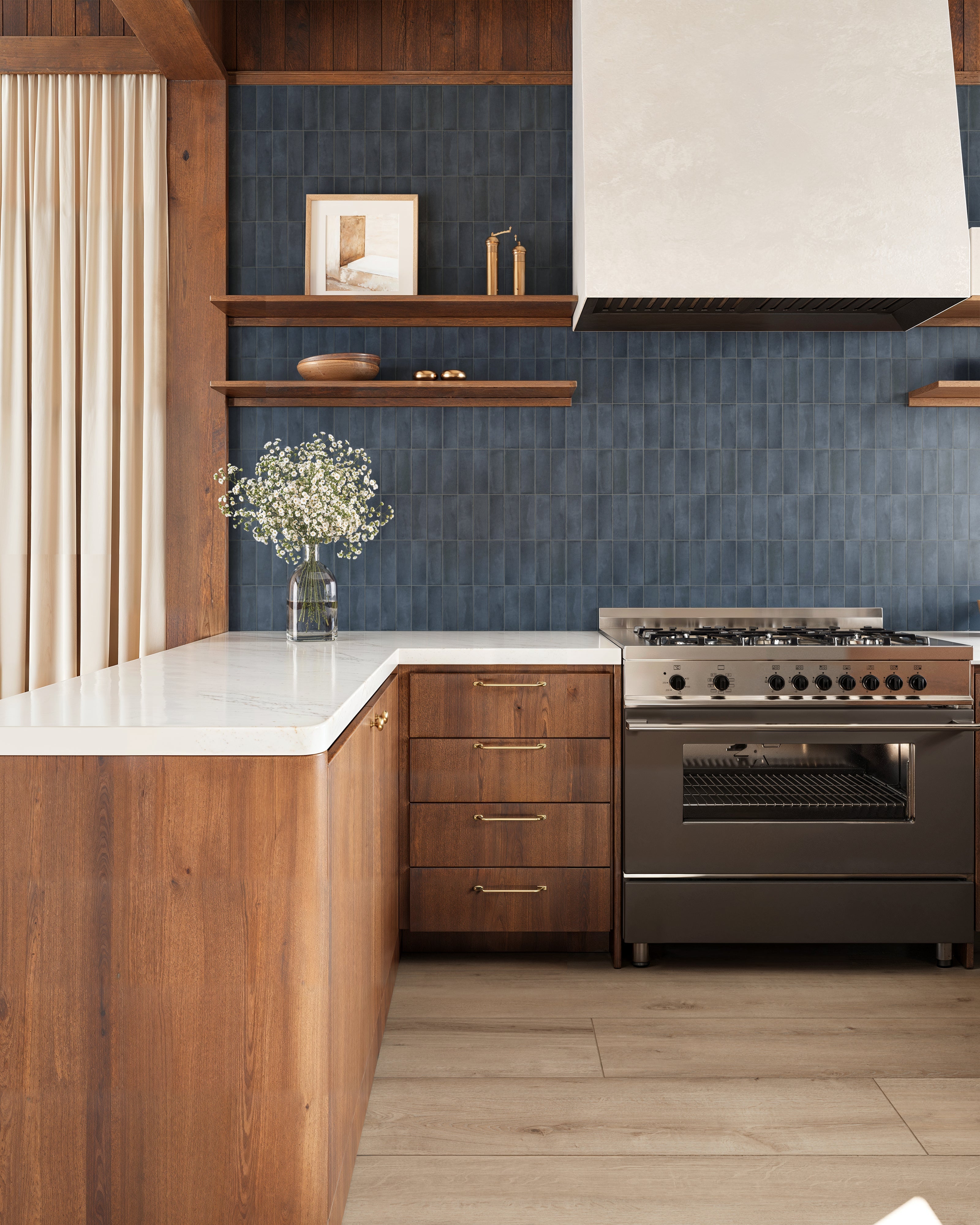 Natasha 2x6 matte porcelain tile in Denim, creating a refined kitchen backsplash with rich navy blue hues, complemented by warm wood cabinets and shelves, accented by a sleek range hood and airy white countertops.