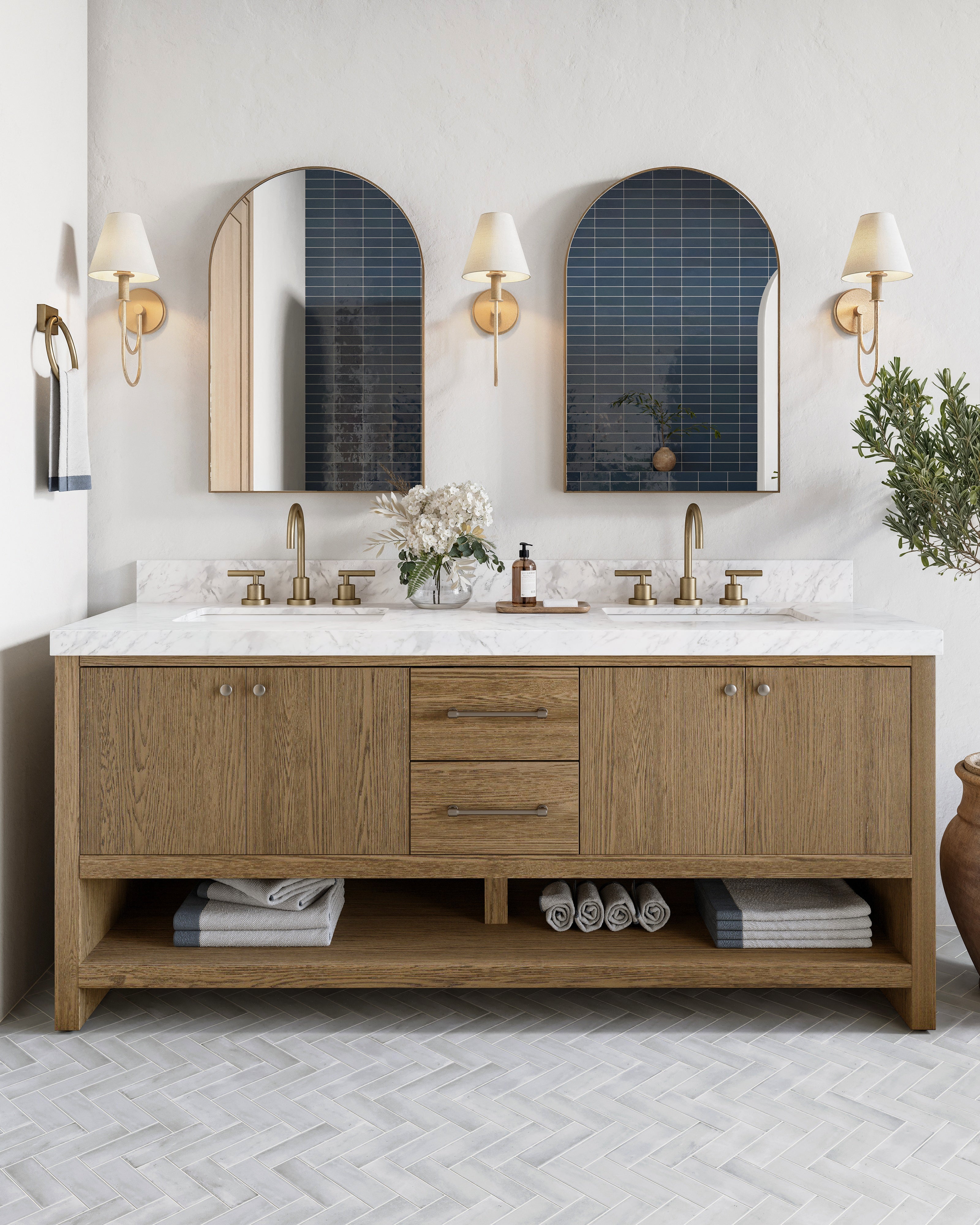 A double vanity bathroom with wood cabinetry, brass fixtures, and arched mirrors reflecting denim blue glossy subway tiles, set above a herringbone tile floor.