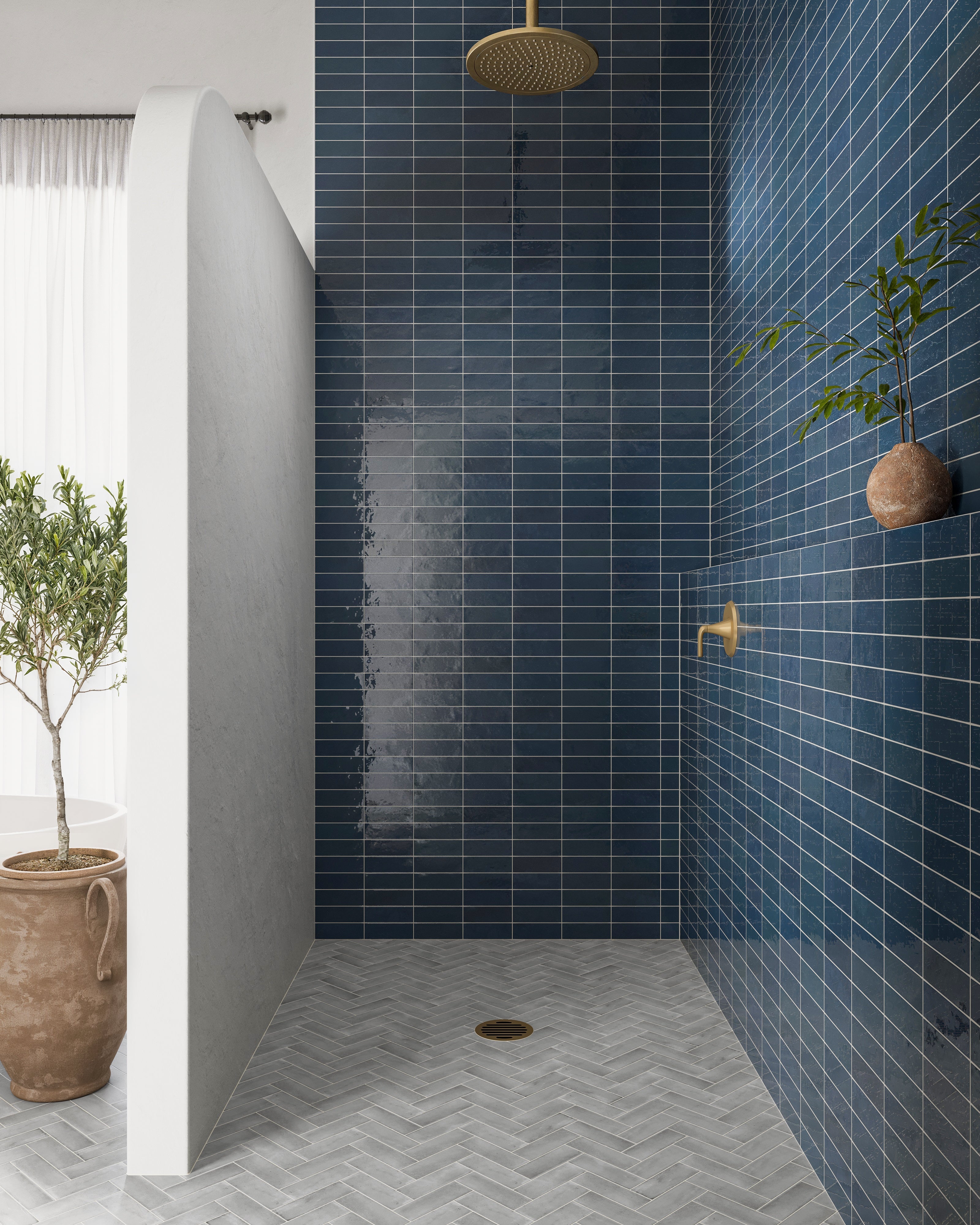 A modern shower clad in glossy dark blue subway tiles with white grout, paired with brass fixtures and a light gray herringbone tile floor.