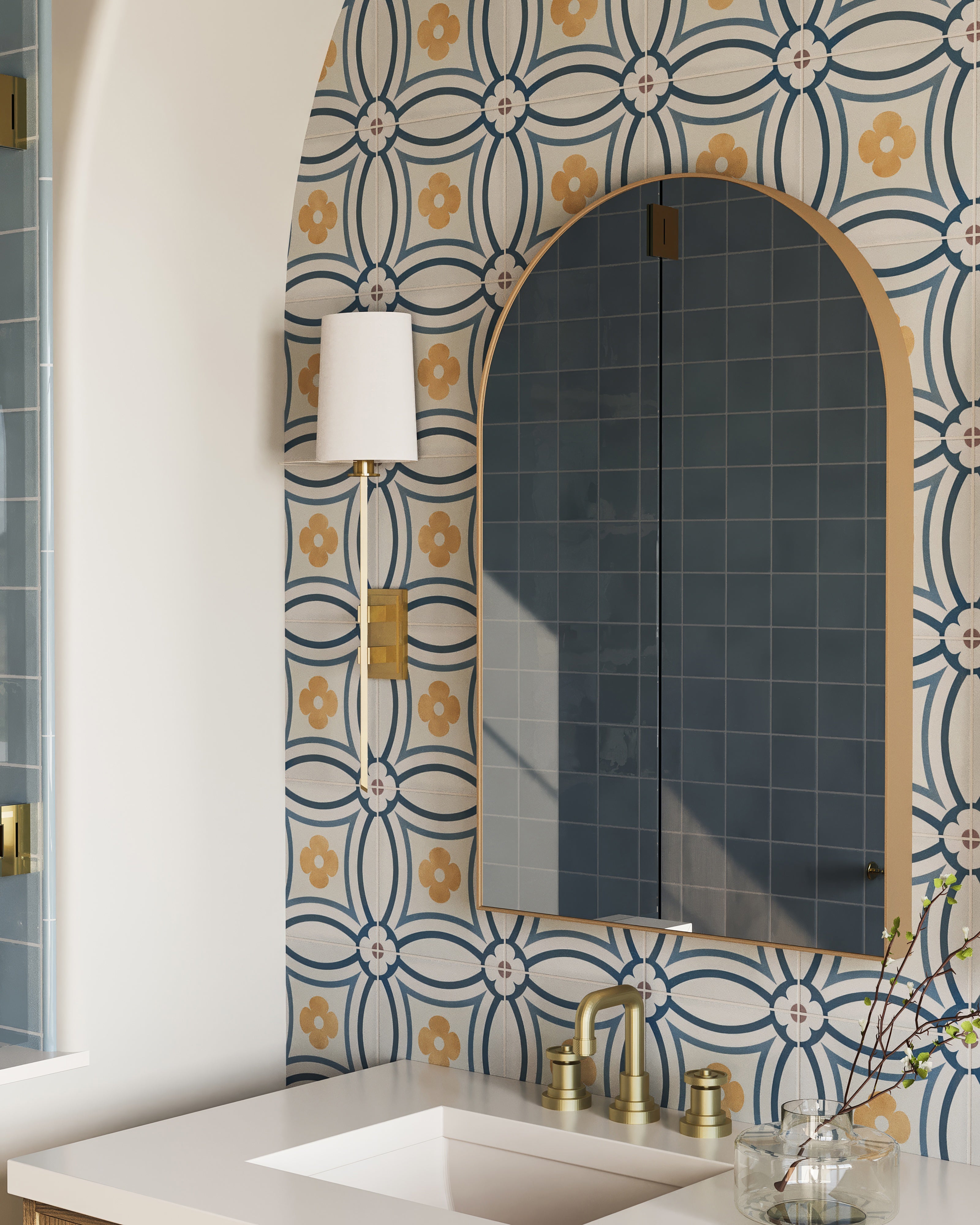 Bathroom with arched brass mirror, floral blue-and-gold tile accent wall, brass faucet, and white countertop with glass vase.